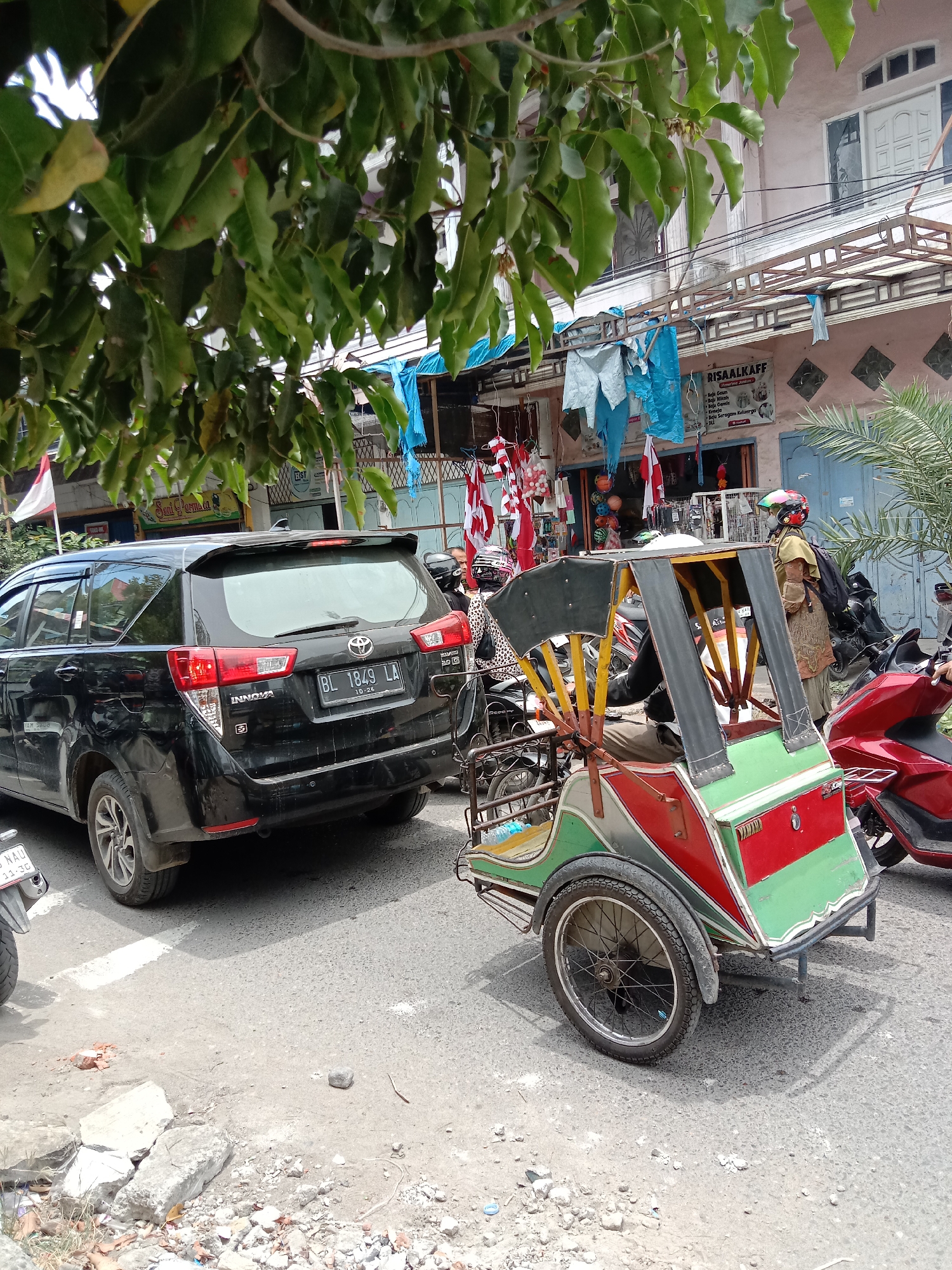 A street scene in Lhokseumawe, Indonesia, features a black SUV, a colorful three-wheeled vehicle, and two individuals. The SUV, identified as an Innova with license plate BL 1849 LA, is parked on the right side of the road. A person wearing a helmet and a patterned shirt is seated in the three-wheeled vehicle, which has red, green, and yellow accents. Another person, partially visible, appears to be standing near the vehicle. Shops with awnings line the street in the background, with laundry hanging on display. A red scooter is parked behind the three-wheeled vehicle. The ground is asphalt, with some debris and cracked concrete visible on the side of the road. Green foliage from a tree partially obscures the upper portion of the image.