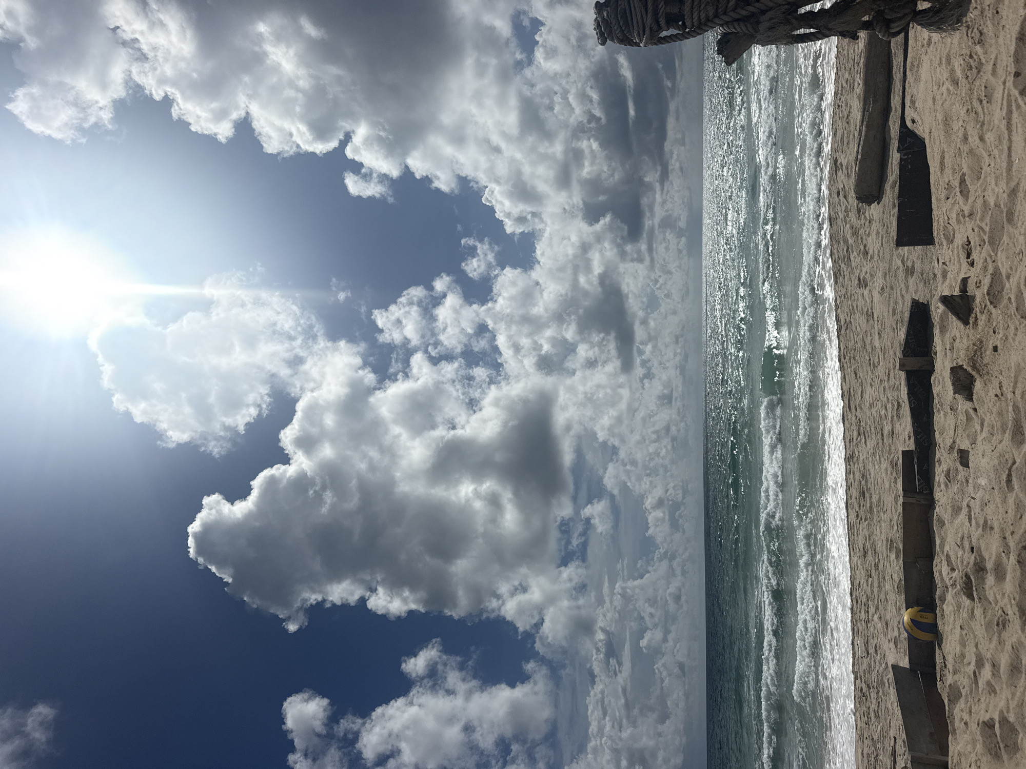 The image depicts a sandy beach scene with the ocean in the background. The sky is a deep blue with scattered white clouds, and the sun is shining brightly, creating a glare on the water. The ocean has moderate waves, and the water is a deep turquoise color. In the foreground, there is a sandy beach with some debris, including pieces of wood and a volleyball. A thick rope is visible on the right side of the frame, attached to a wooden post. The overall impression is a bright, sunny day at the beach, likely in a tropical location given the vibrant colors of the water and sky. The specific location in Choeng Thale, Thailand, is not evident from the image itself, but it is described as a beach setting.