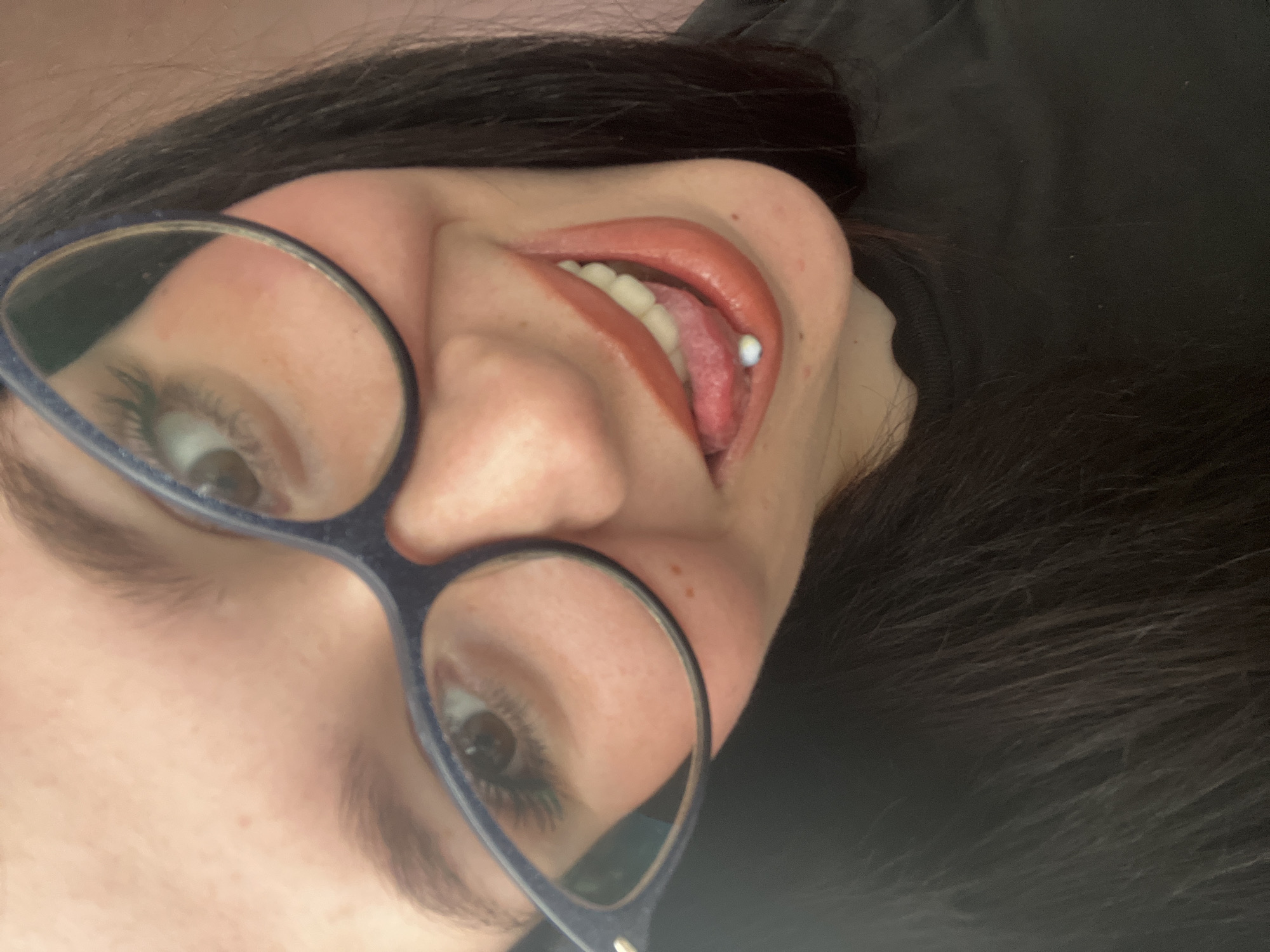 The media file is a close-up, vertically oriented photograph of a person's face, primarily focusing on their mouth and eyes. The person is wearing dark-rimmed glasses. Their mouth is slightly open, revealing teeth and a tongue with a tongue piercing that has a small, light-colored bead. The person has dark hair. The background is dark and out of focus, suggesting a dimly lit indoor setting. There are no visible text, specific time of day, or weather cues. The scene is intimate and personal, with the individual looking towards the camera. The facial expression is neutral or slightly playful due to the open mouth and tongue presentation.