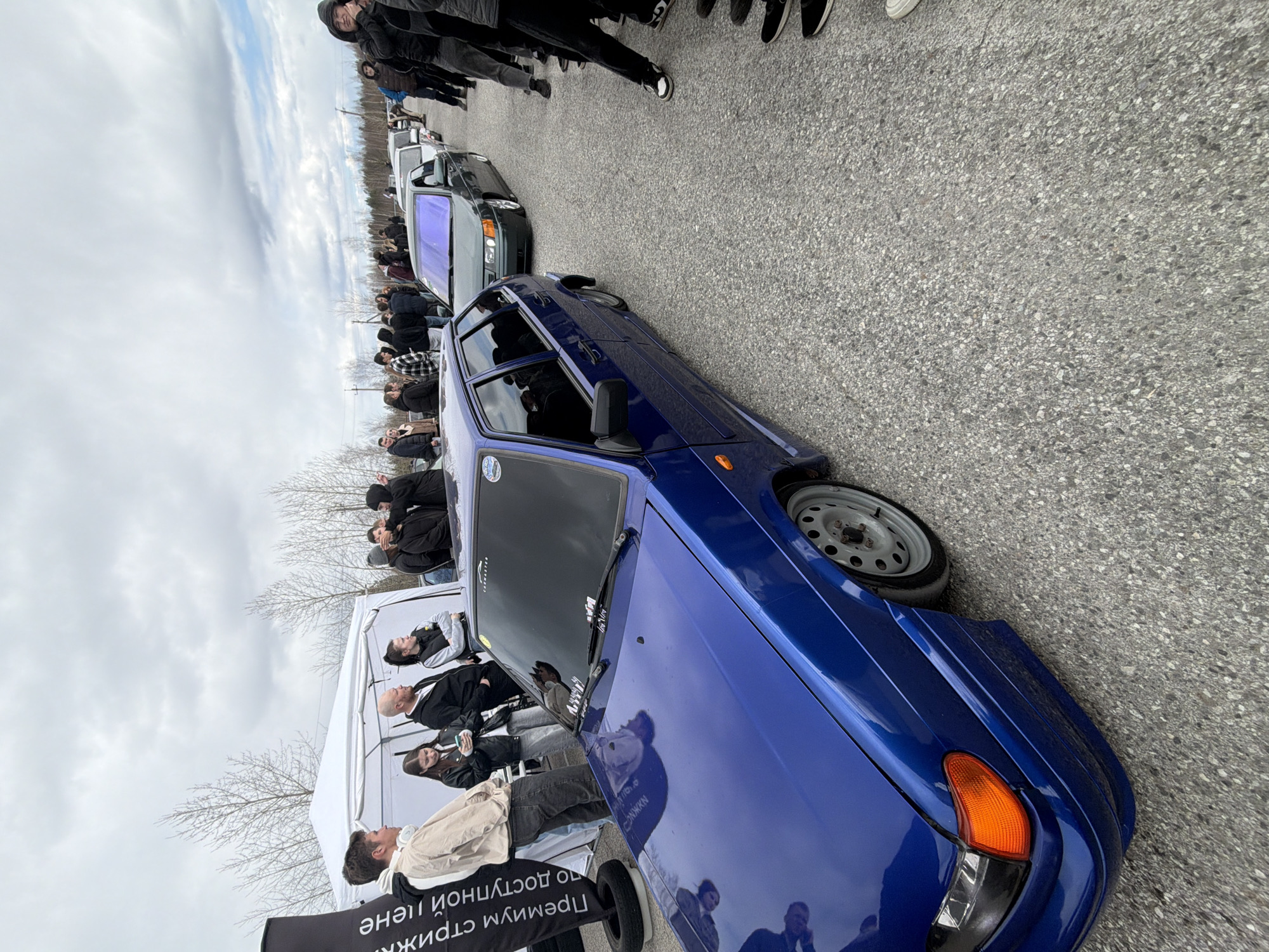 The image shows a street in Perm, Russia with a line of cars parked on the side of the road. People are gathered around the vehicles, some under a tent. A blue car is prominent in the foreground. The sky is overcast.