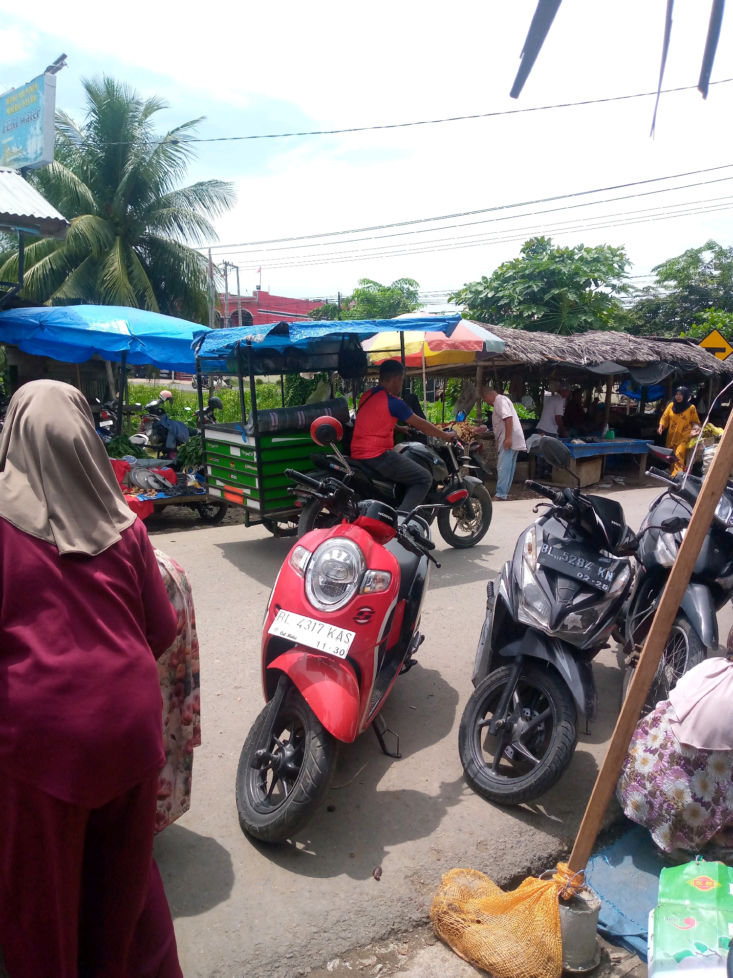 The image captures a street scene in Keudeteupinpunti, Indonesia, likely a market or roadside trading area. In the foreground, a person wearing a burgundy hijab and dress stands facing away from the camera, their body partially obscuring a red scooter. To the right, a black scooter with license plate "BL 5284 KN" is parked. A wooden pole leans against a gray object, and an orange mesh bag lies on the ground near it. In the background, several market stalls are visible, covered with blue and thatched roofs. People are seen interacting at these stalls, with one individual in a red shirt riding a motorcycle and another in a white shirt attending to goods. A green utility cart is also present, laden with items. The environment appears sunny with a clear sky and some trees and power lines visible.