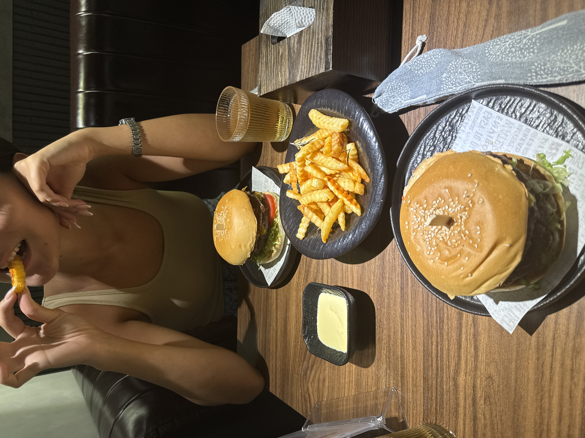 An overhead shot shows a woman eating a French fry at a restaurant table. The table is laden with food including two burgers, a plate of French fries, and a small dish of mayonnaise or aioli. A glass of liquid, possibly beer or iced tea, is also visible. The restaurant appears to be dimly lit and modern in design. It's impossible to determine the location with certainty in Đà Nẵng, Vietnam, but the style of food and restaurant suggests an upscale establishment.