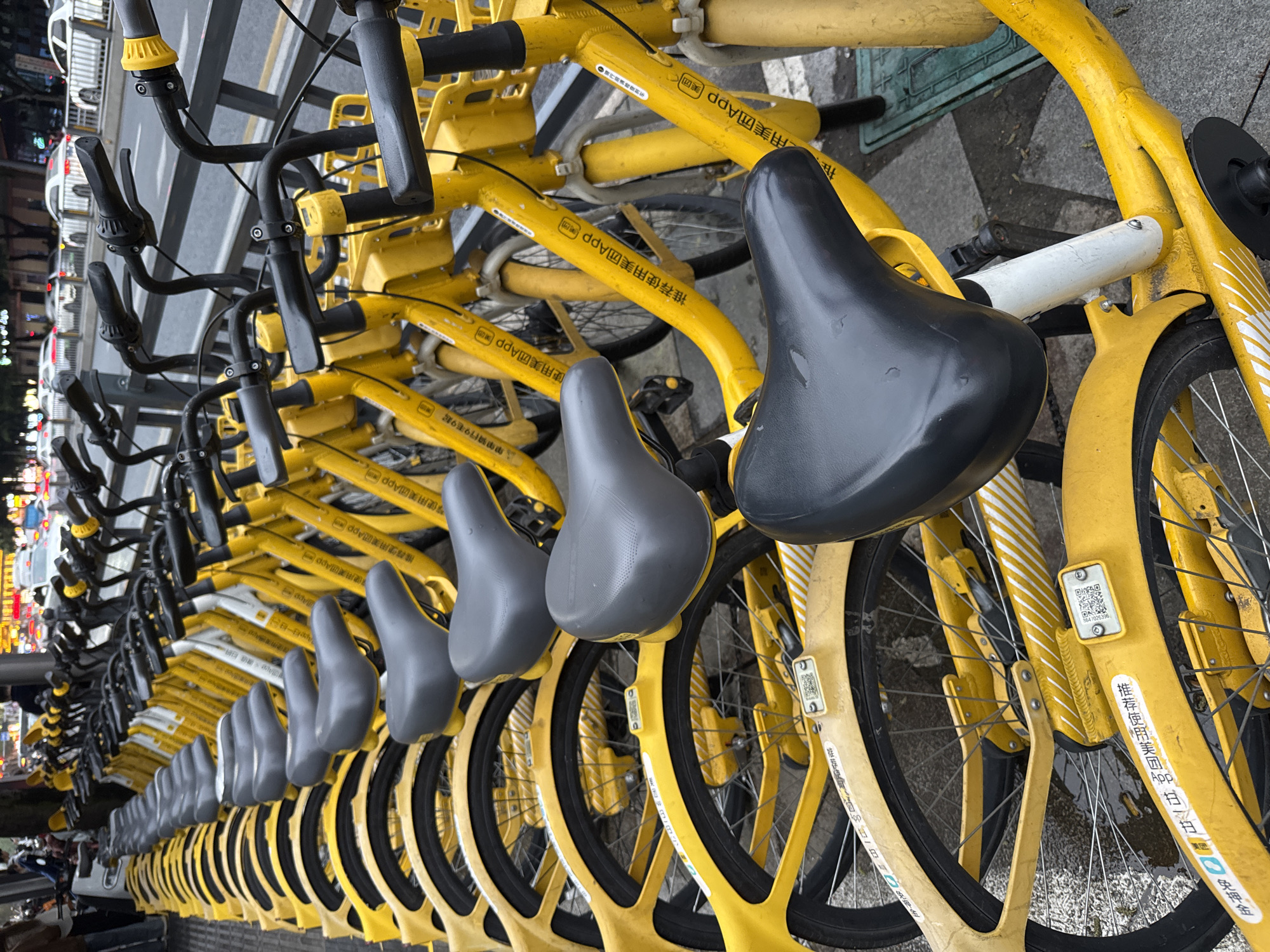 A row of yellow shared bicycles, parked on a city street in Jinjiang District, China, fills the frame. The bicycles are closely aligned, with their black seats and handlebars creating a repeating pattern. The background reveals glimpses of urban life, including buildings, streetlights, and pedestrians. The pavement is wet, reflecting the city lights. The bikes have Chinese text in yellow along the frames.