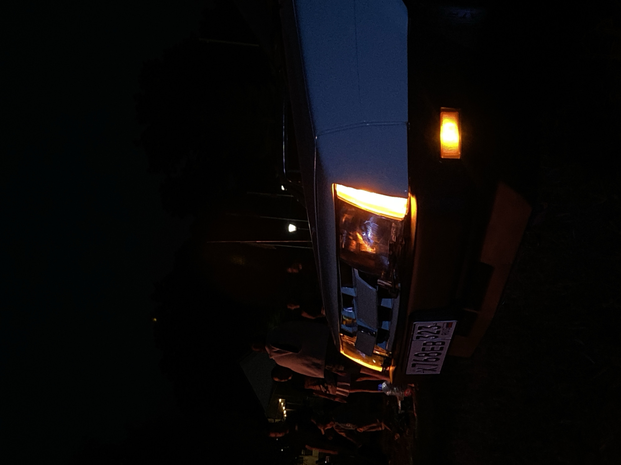 In Bezymyannoe, Russia, at night, a close-up shot captures the front of a light-blue car with its headlight illuminated. The headlight is a bright orange, U-shaped light. Below it, a small rectangular orange marker light is also lit. The car's license plate, visible at the bottom left, displays the characters "X768EB 323". In the background, several people are standing near a tent, their silhouettes indistinct in the darkness. The scene suggests a gathering or event taking place outdoors at night.