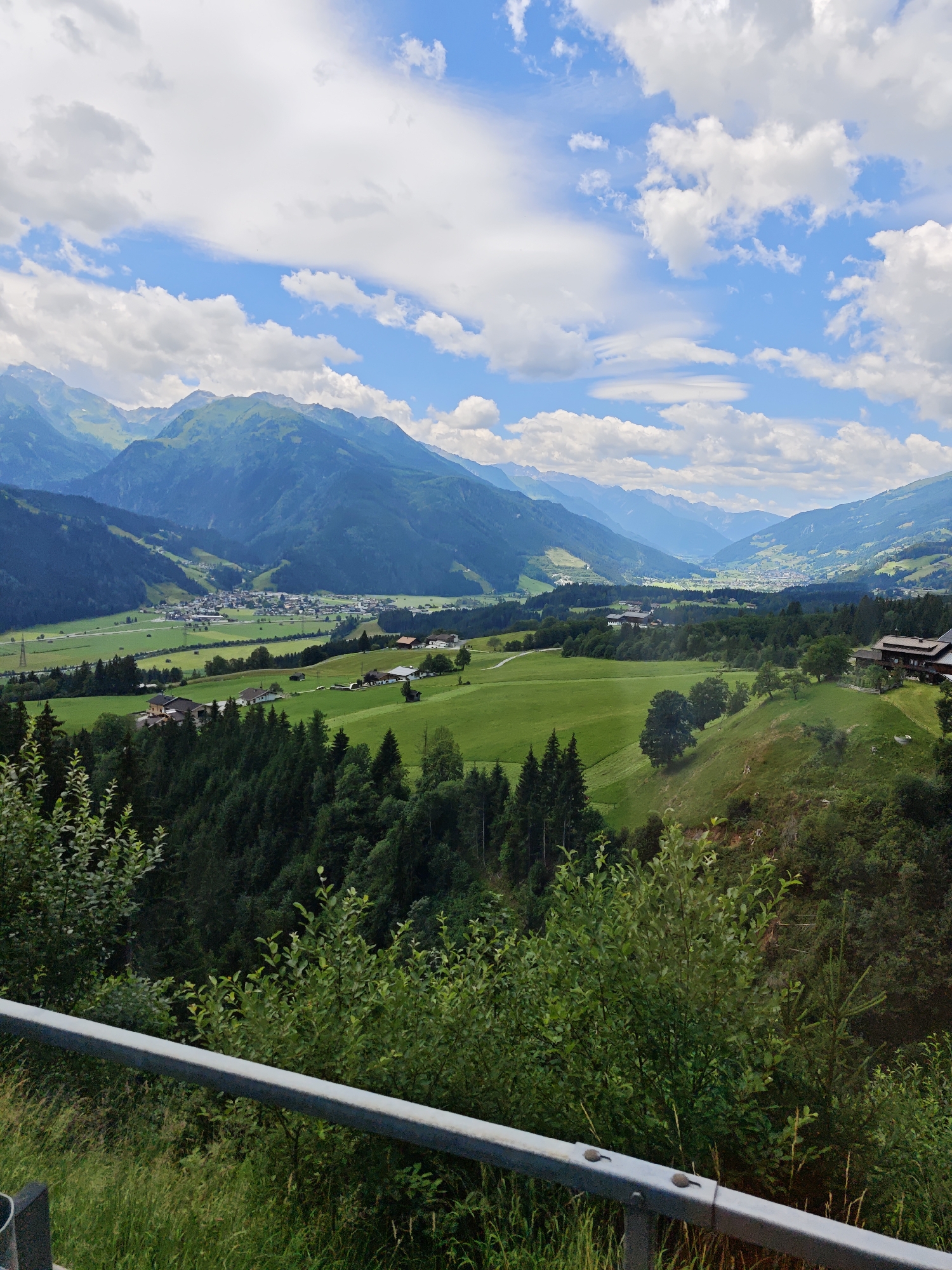 This is a medium-format, eye-level, full outdoor shot of a mountain valley in Mittersill, Austria. In the foreground, a metal guardrail runs horizontally across the bottom of the frame, with lush green vegetation to its right and in the lower left corner. Behind the guardrail, the scene opens up to a vast expanse of rolling green hills dotted with trees and scattered farmhouses. In the midground, a dense forest lines the base of the mountains, which rise majestically in the background. The mountains are covered in a patchwork of green forests and rocky outcrops, with hints of snow near the peaks. The sky is a brilliant blue, filled with fluffy white clouds. A small village or town is nestled in the valley in the distance. The overall impression is one of serene natural beauty and rural tranquility.