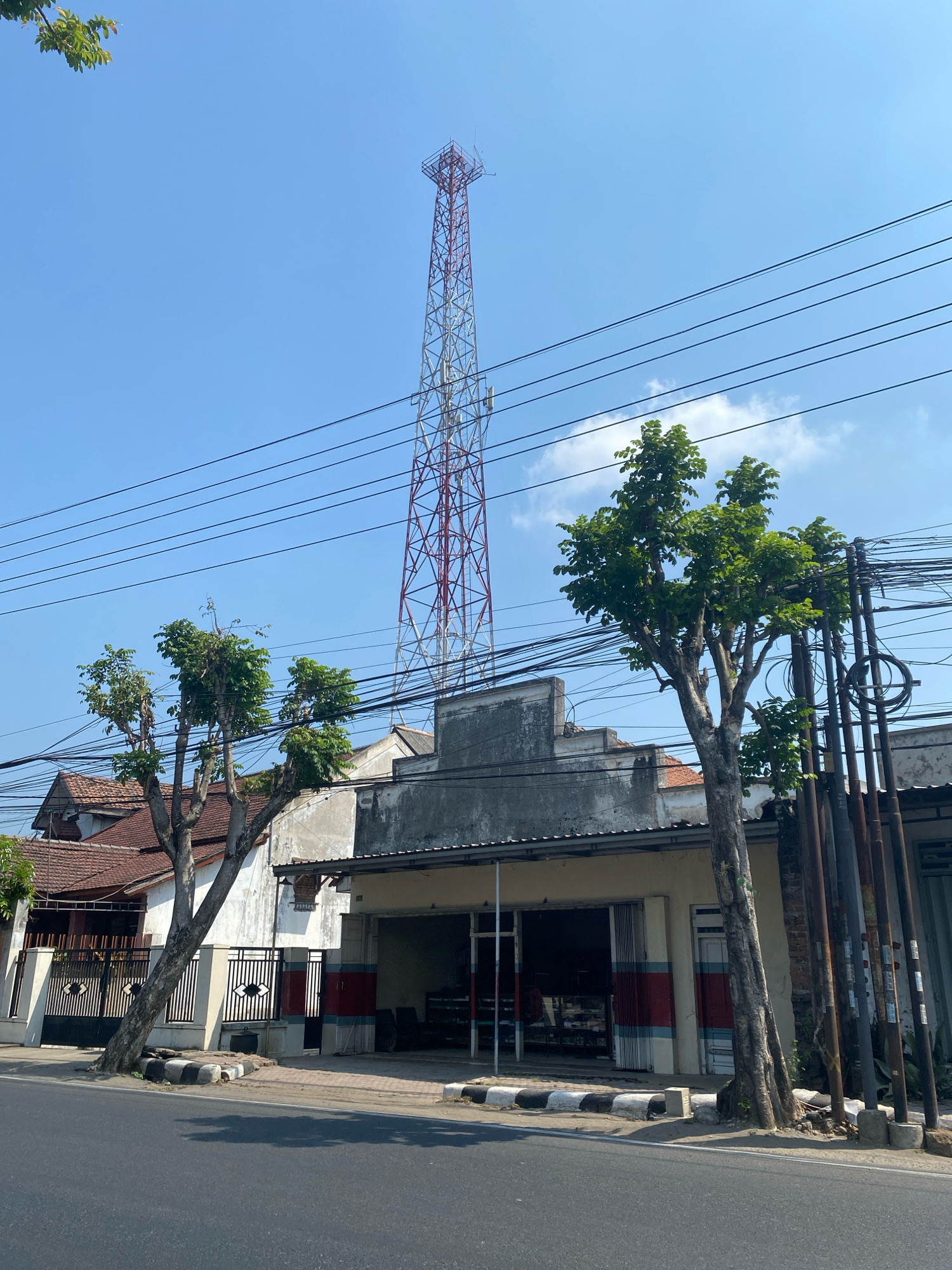 The image depicts a street scene in Kediri City, Indonesia, on a clear day with a bright blue sky. Dominating the background is a tall, red and white lattice telecommunications tower. In the middle ground, several buildings line the street. One prominent building appears to be a shop with an open-fronted entrance displaying shelves and goods inside. The shop has a faded facade with red and blue stripes. To the left of this shop, a residential building with a tiled roof is visible behind a fence. Several mature trees with lush green foliage are scattered throughout the scene, some appearing pruned. A network of overhead power lines crisscrosses the sky, supported by poles. The foreground shows a paved road and sidewalk with concrete barriers and some vegetation. The overall impression is of a typical urban street with a mix of commercial and residential structures, characteristic of a Southeast Asian city. There are no people or animals visible in the image.