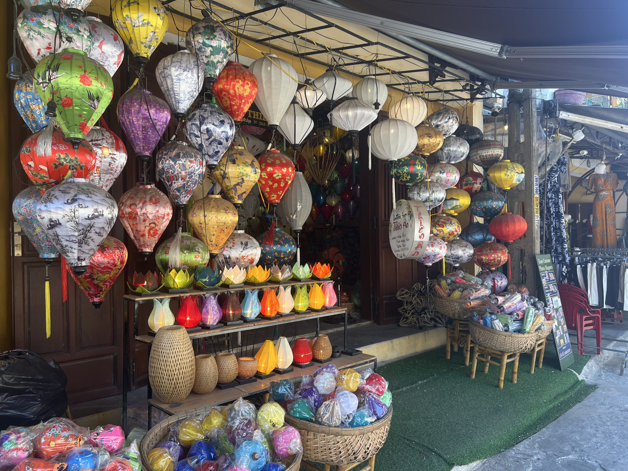 A storefront in Hoi An Ward, Vietnam, displays an abundance of colorful silk lanterns. The lanterns, varying in size, shape, and design, are hung from the ceiling and displayed on shelves and in baskets. Many lanterns are patterned with floral motifs, birds, and scenic landscapes. The scene captures a vibrant street market atmosphere.