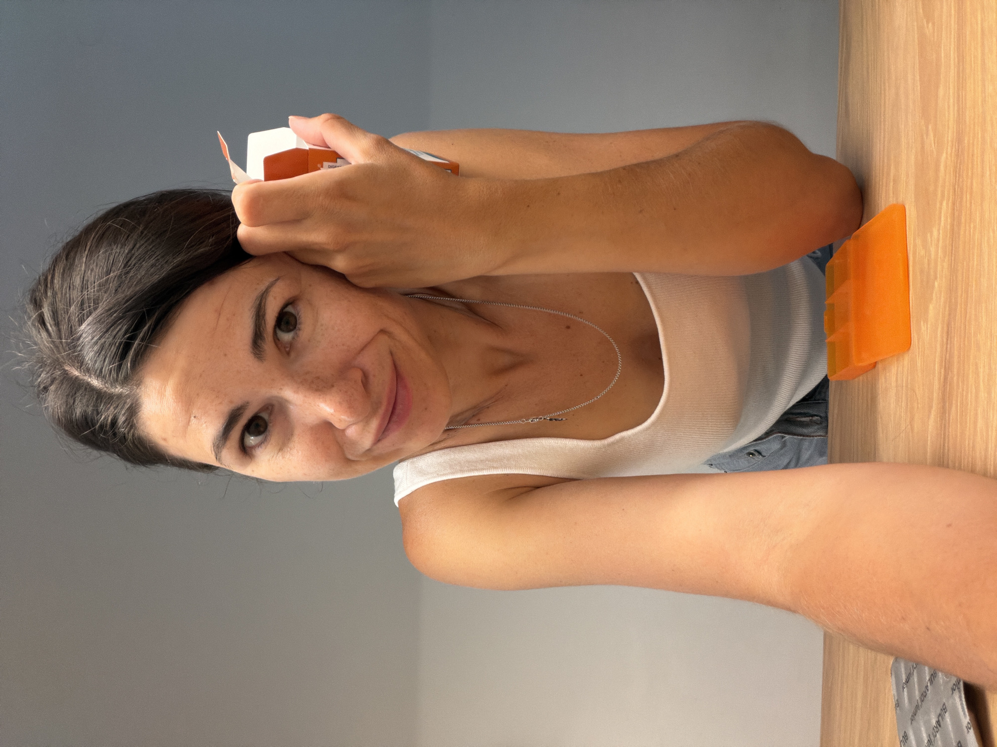 A young woman with dark hair and freckles is looking at the camera and holding a small orange and white box to the side of her head. She is wearing a white tank top and a silver necklace. She is leaning forward over a wooden surface. To her right, on the wooden surface, is an orange plastic pill organizer. To her left, also on the wooden surface, is a silver blister pack. The background is a plain gray wall. The scene appears to be indoors, possibly a home or office. There is no indication of the city of Budva, Montenegro.