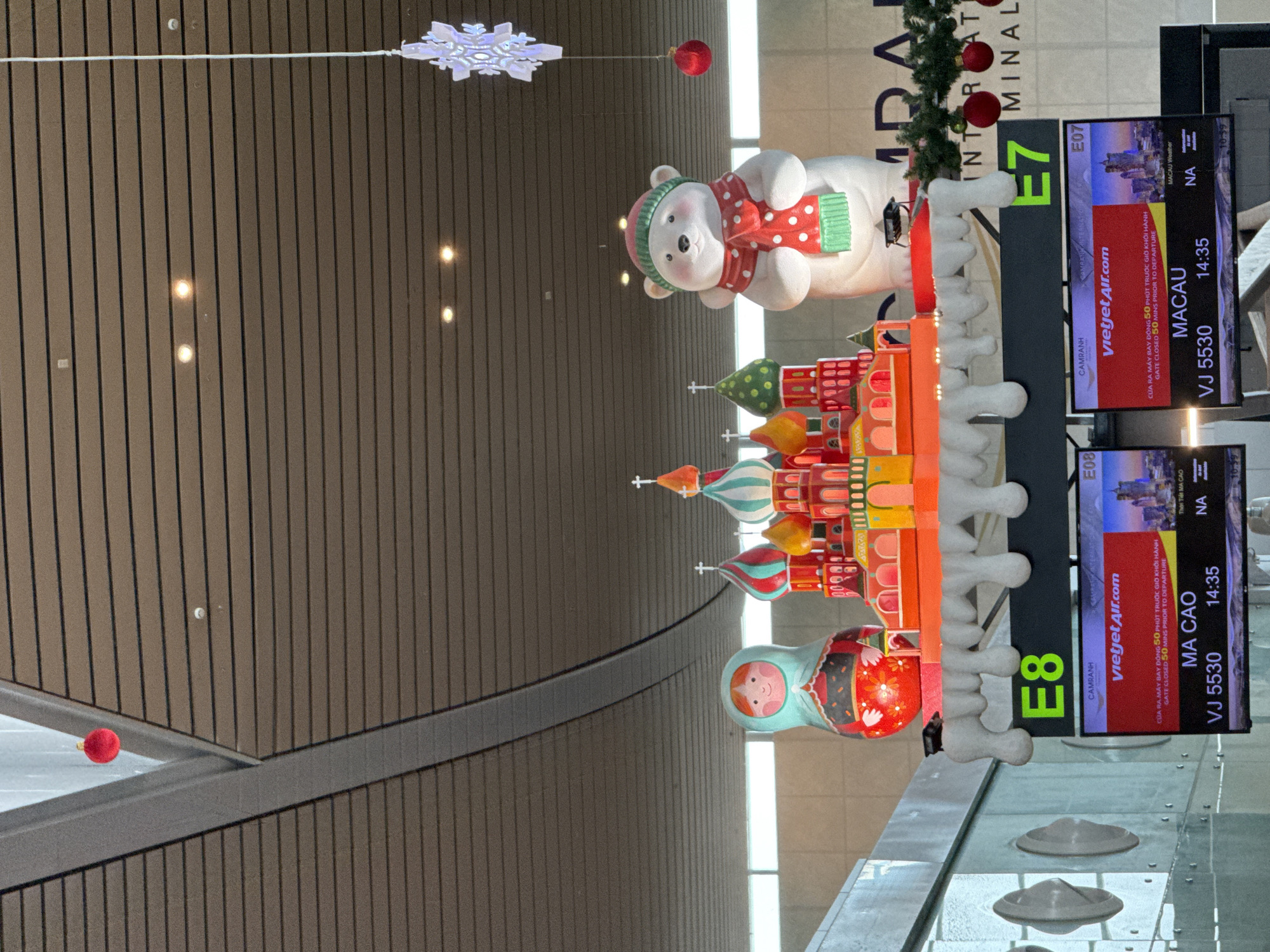 The image shows a festive airport departure hall decorated for the holidays. In the foreground, an electronic display board shows flight information. One segment of the board displays "MA CAO" with flight number "VJ 5530" and departure time "14:35", indicating a flight to Macau. Another segment shows "MACAU" with flight number "VJ 5530" and departure time "14:35", also for Macau. The display board is positioned below a large, colorful Christmas display featuring a Russian nesting doll, a model of Saint Basil's Cathedral, and a polar bear wearing a Santa hat and scarf. This display is adorned with artificial icicles and Christmas ornaments. In the background, a partial view of the "CAMRANH INTERNATIONAL AIRPORT" signage is visible, confirming the location as Cam Ranh, Vietnam. The ceiling of the airport is made of dark horizontal planks, with modern lighting fixtures. Red spherical ornaments and a snowflake-shaped light fixture hang from the ceiling, contributing to the holiday atmosphere.