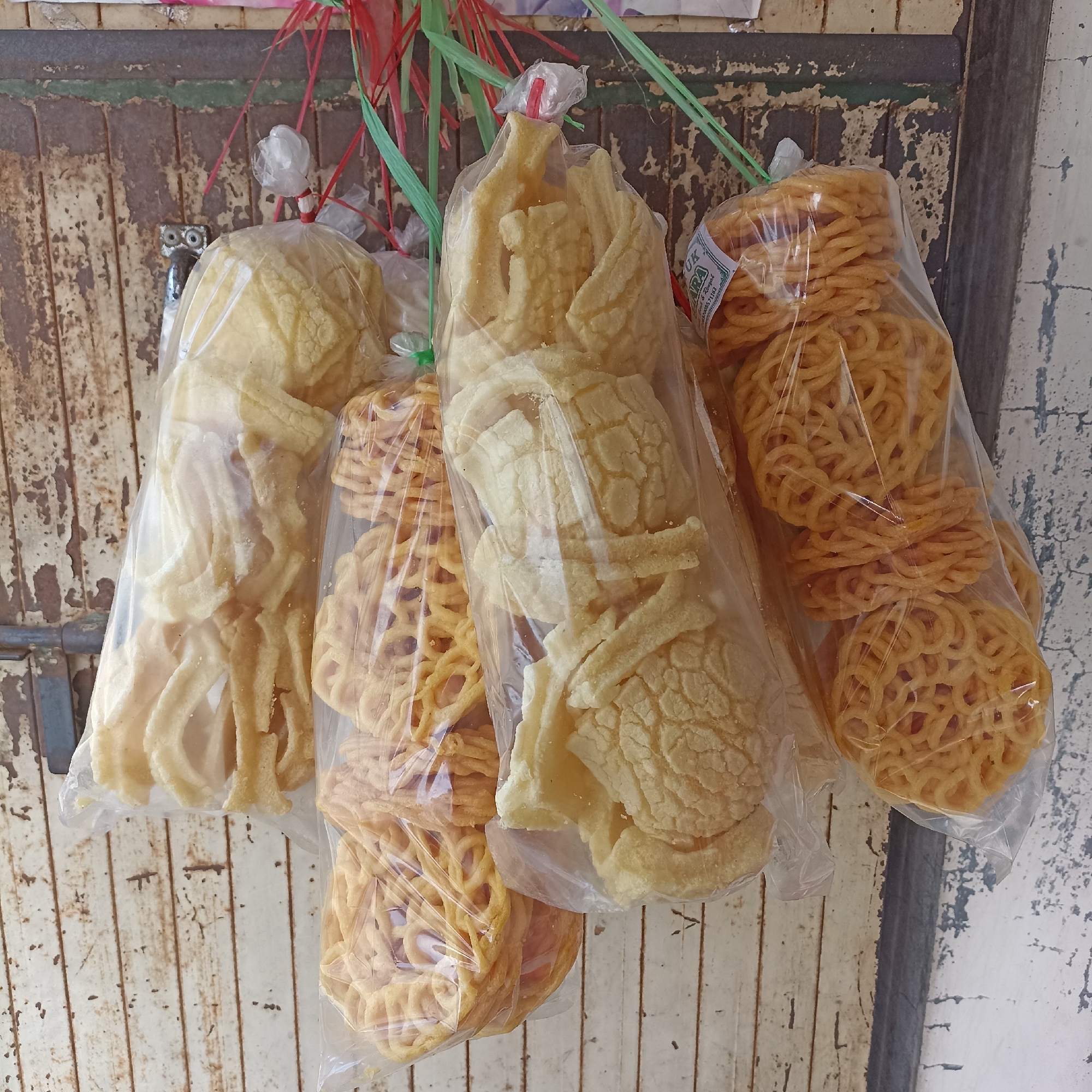 Four plastic bags of snacks are hanging from a weathered wooden surface. The snacks appear to be a type of cracker or chip. From left to right, the first bag contains pale yellow, irregularly shaped chips. The second bag contains round, intricately patterned, golden-brown crackers. The third bag contains a similar type of pale yellow, irregularly shaped chips as the first bag, but these appear to be larger. The fourth bag contains more of the round, intricately patterned, golden-brown crackers, some of which have a label attached. The wooden surface behind the snacks is painted white and appears to be the door or wall of a shop. The overall scene suggests a street vendor stall in Magelang, Indonesia.
