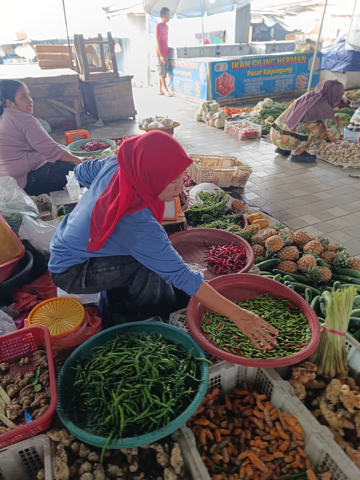 In Kayu Agung, Indonesia, a market stall displays a variety of fresh produce. A woman wearing a red hijab and a blue shirt is attending to a large bowl of green chilies. Nearby, another woman in a light purple shirt is seated. In the background, various vegetables like pineapples, cucumbers, and ginger are arranged for sale. A sign with "IKAN GILING HERMAN PASAR KAYUAGUNG" is visible, indicating the market's name and location. The scene captures a moment of commerce within a bustling marketplace, highlighting the daily activities of local vendors and shoppers.