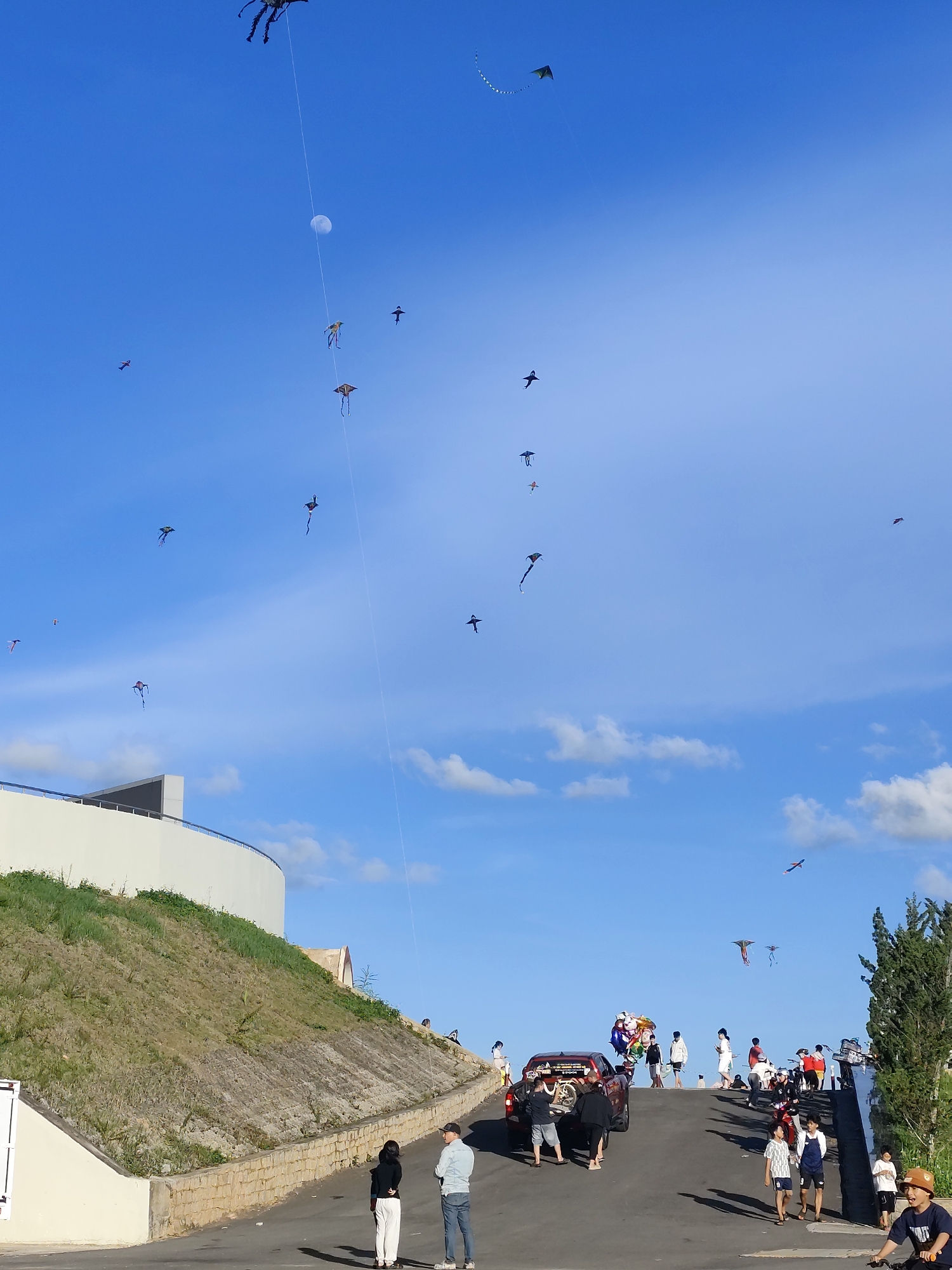 A group of people are gathered on a paved incline under a clear blue sky, likely for a kite-flying event. Many kites are visible in the sky, in various shapes and colors, with some appearing to be complex designs. A prominent kite, resembling a large spider, is tethered to the ground and dominates the upper left portion of the frame. The moon is also visible in the sky.
The scene depicts a daytime event, with bright sunlight casting shadows on the ground, suggesting it is mid-morning or afternoon. The weather appears to be fair and clear, with a few wispy clouds scattered across the sky. The setting includes a grassy slope and a modern-looking white building with a railing on the left side of the image. On the right, there are trees and more people gathered, some with bicycles and motorcycles.
A red pickup truck is parked on the incline, adorned with colorful balloons and possibly decorations related to the event. Several individuals are scattered across the scene, some standing and watching the kites, others walking or interacting. There are also children present, adding to the festive atmosphere. The overall mood appears to be relaxed and celebratory.
While no specific text is clearly legible in the image, the context suggests a local festival or recreational gathering, possibly in Da Lat, Vietnam, as indicated by the location context. The diverse group of people and the presence of traditional and modern kites hint at a cultural or community event.