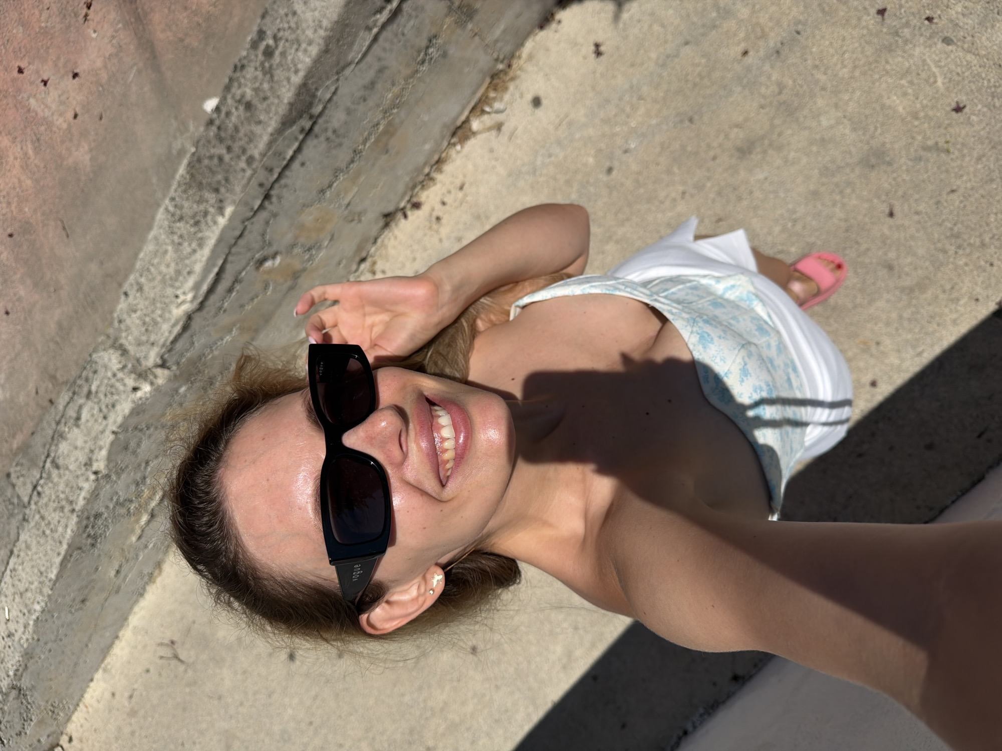 A young Caucasian woman with blonde hair is taking a selfie outdoors, with a concrete structure and a textured ground visible. She is wearing black sunglasses with "VOGUE" written on them and a strapless white top with a blue floral pattern. Her right hand is adjusting her sunglasses, and she has a small stud earring with a dangling charm in her left ear. Her expression is a bright smile, revealing her teeth. She is wearing a pink flip-flop on her right foot. The lighting suggests it is a sunny day, casting shadows on her arm and the ground. The background appears to be an outdoor staircase or elevated platform, possibly in a public space in Budva, Montenegro, given the style of the concrete and stonework.