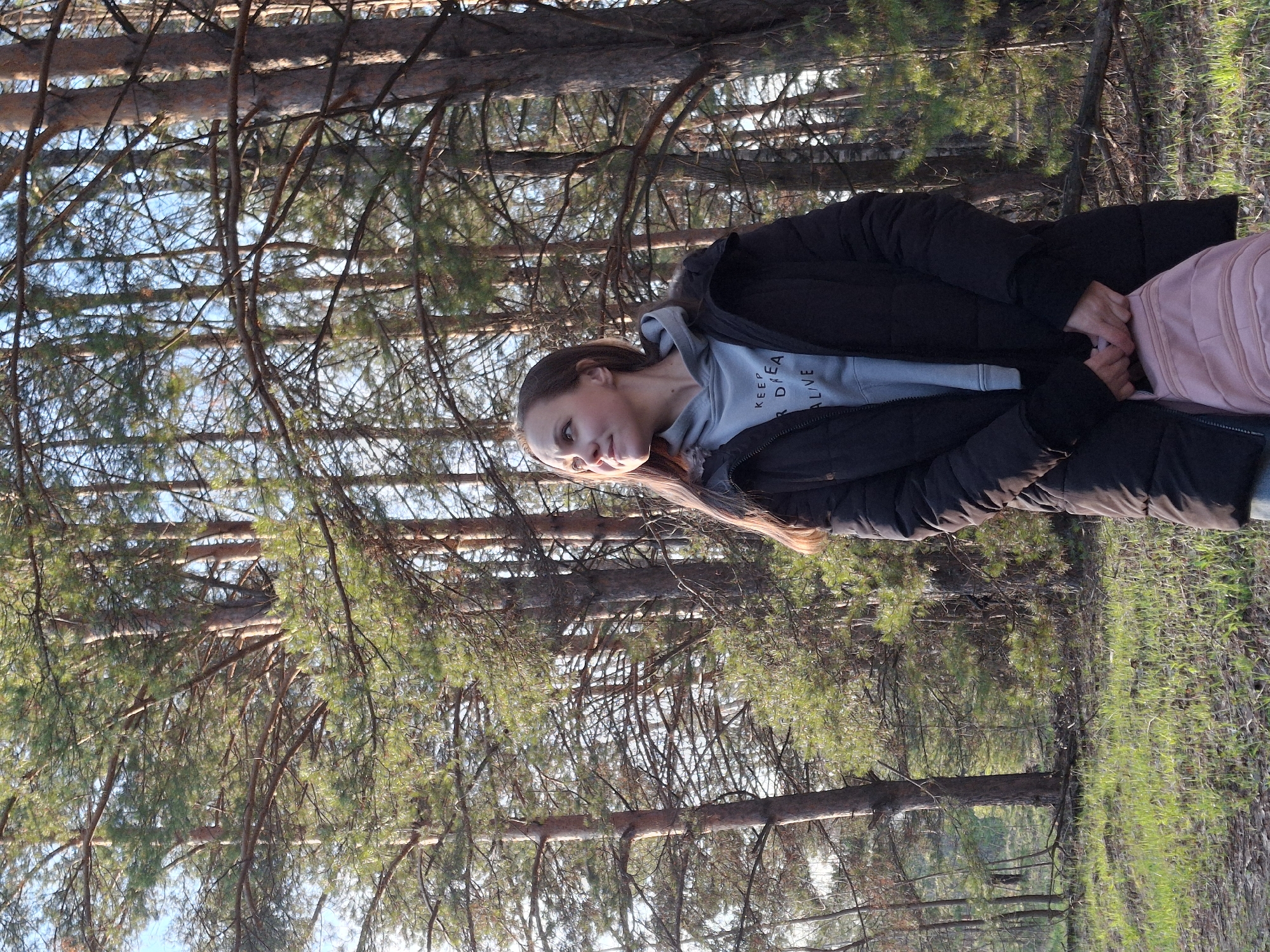 A young woman is depicted standing in a pine forest, with the original image oriented sideways. She is positioned towards the center-right, looking slightly off-camera with a neutral and reflective expression. Her long, light brown hair cascades over her shoulders. She wears a black puffer jacket over a light blue hooded sweatshirt. Partially visible text on her hoodie reads "KEEP CA / R DREA / ALIVE," likely signifying "KEEP YOUR DREAMS ALIVE." Her hands are clasped in front of her, holding onto a light pink item, possibly a backpack strap or part of her attire.
The setting is a dense pine forest, characterized by numerous tall trees with reddish-brown trunks and green needles extending throughout the frame. The tree branches interlace to form a canopy, through which patches of clear blue sky are visible. The forest floor shows a scattering of green grass and sparse undergrowth. The lighting appears soft and warm, suggesting either early morning or late afternoon. The scene conveys a tranquil, natural environment, consistent with a forest in Chaikovsky, Russia.