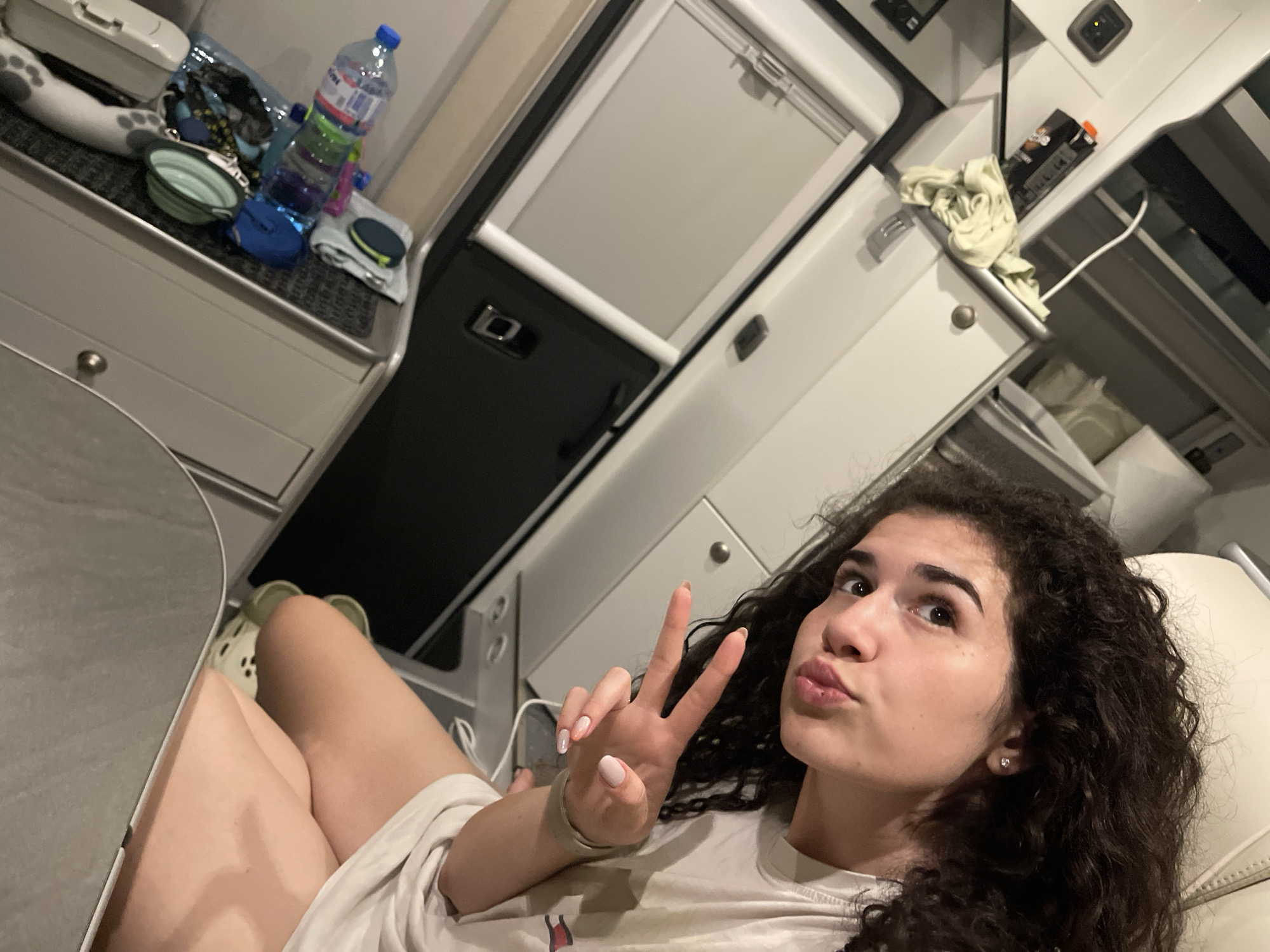 An image, rotated ninety degrees clockwise, displays an indoor scene in Koprinka, Bulgaria, featuring a young adult female reclining on a light-colored surface. The female subject has dark, curly hair and is wearing a light-colored t-shirt. Her right hand is raised, forming a peace sign, displaying light pink nail polish on her manicured fingernails. Her lips are slightly puckered in a pout. She wears light-colored (possibly pale green or beige) Crocs on her feet, which are partially obscured by a light-colored table or shelf in the upper left of the frame.
The environment appears to be a compact, modular interior space, characteristic of a camper van, RV, or small cabin, constructed with light gray and white paneling and surfaces. In the upper right quadrant of the image, resting on a gray carpeted surface within an open compartment or drawer, are several objects: a green collapsible bowl, a blue container, a clear plastic water bottle with colorful bands ("DEVIN" label partially visible), and other small, indistinct items.
Below the female subject's head, a light-colored cabinet or wall panel is visible. To the right of the subject, a black rectangular object with a silver handle is mounted on a white surface, next to a window or vent equipped with a white pull-down blind. Further down the right side, another open compartment reveals a crumpled, light-green cloth, a white electrical cable, and a black rectangular box. The overall lighting is consistent with an indoor setting, likely artificial.