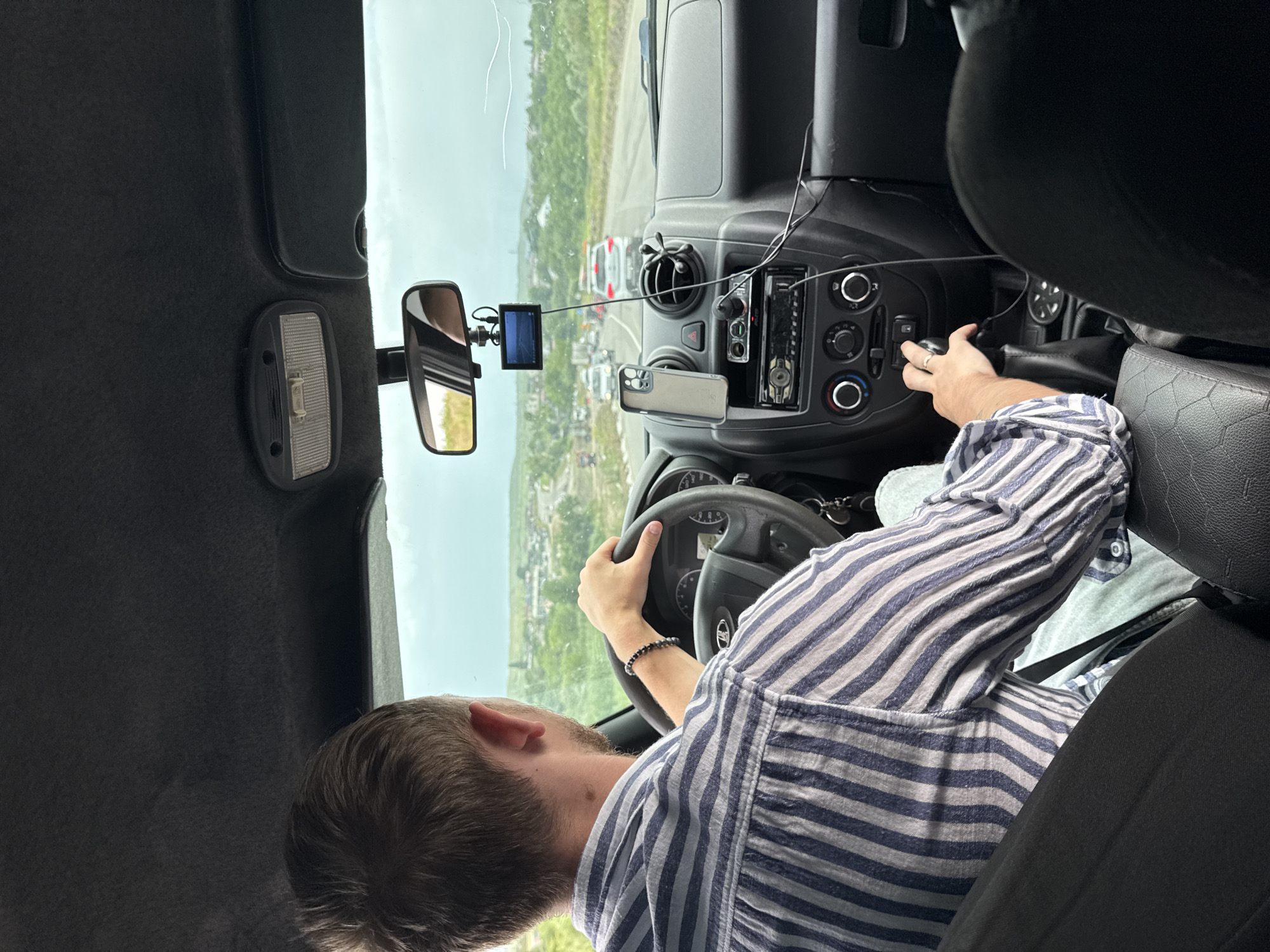 The image shows the interior of a car, looking forward from the back seat. A man with short brown hair is driving, wearing a blue and white striped shirt. He is holding the steering wheel with his right hand and has a black beaded bracelet on his left wrist. In front of him, the dashboard is visible, with a car stereo, air vents, and a small screen mounted on the windshield. A cell phone is attached to the dashboard. Outside the car, a road is visible with other vehicles and a rural landscape with buildings in the distance. The sky is overcast. The scene appears to be in the city of Nadezhda, Russia.