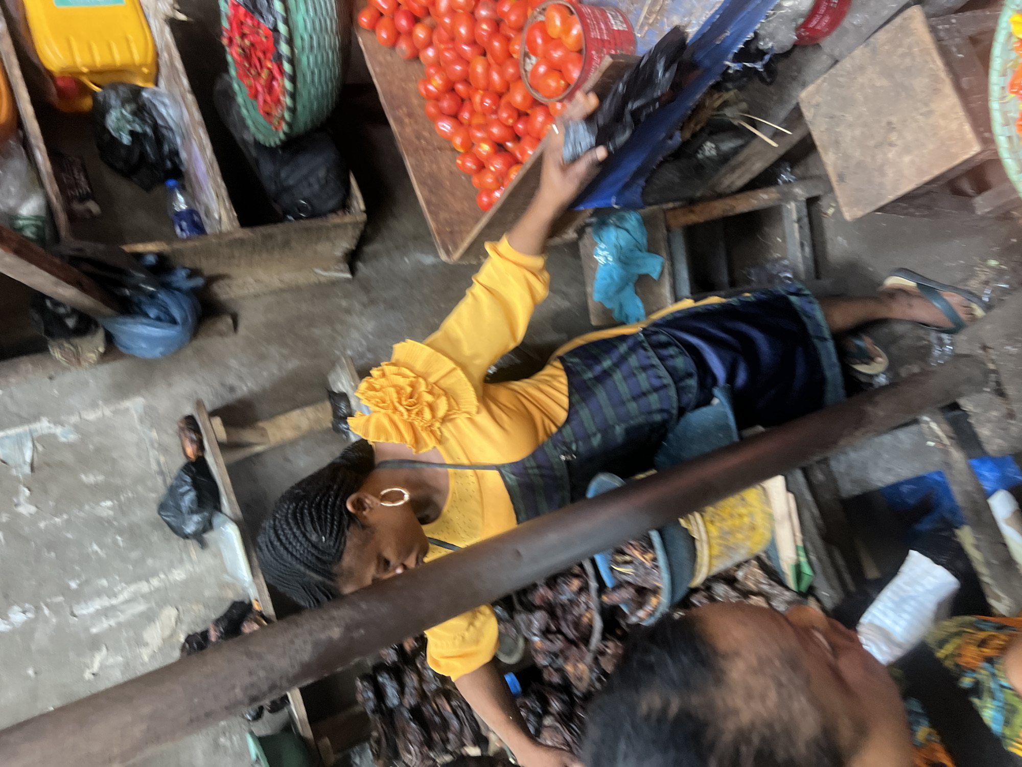 In Nkpor, Nigeria, within a market stall, a Black woman with braided hair wearing a bright yellow top and a blue and green checkered apron is attending to her produce. She is surrounded by various goods, including what appears to be dried fish in buckets and baskets, and a large display of red tomatoes in wooden crates. The stall is dimly lit, with rough-hewn wooden shelves and concrete walls visible. The woman is in the process of handling some black objects, possibly more produce or packaging materials, and her posture suggests she is engaged in a transaction or preparation. Another person, partially visible in the foreground, is also present in the stall, though their actions are not entirely clear. The overall scene depicts a vibrant market environment, typical of such locations in Nigeria.