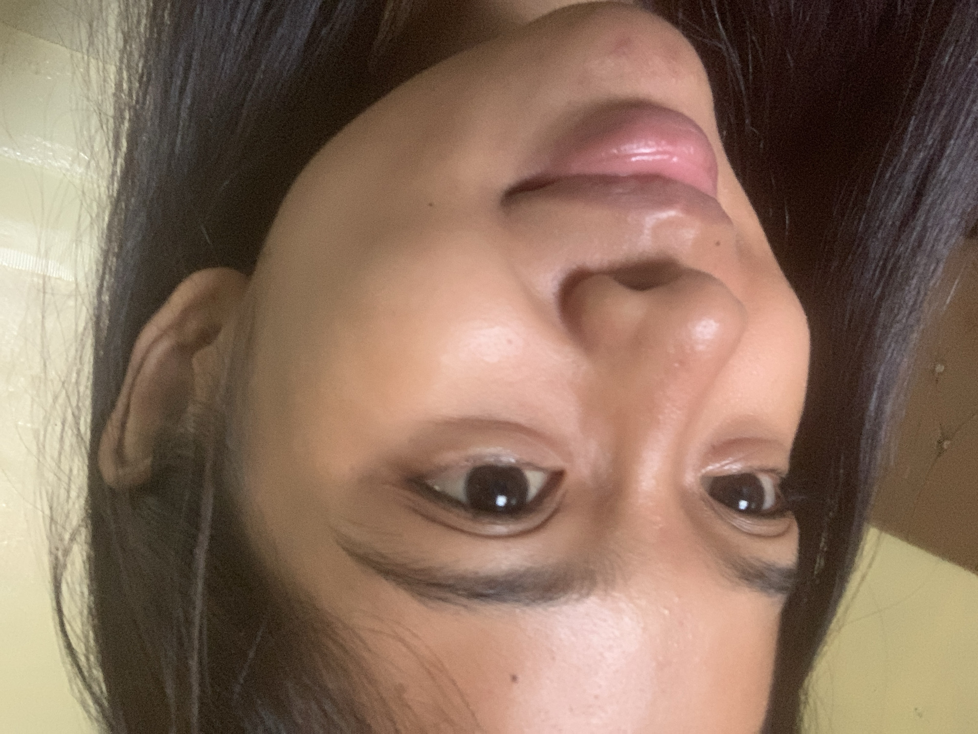 A close-up, head-and-shoulders shot shows the right side of a young woman's face. She has dark hair, brown eyes, and medium-toned skin. The lighting is warm and somewhat diffused, creating a slight sheen on her skin. Her lips are slightly parted, revealing a hint of teeth. The background is out of focus and appears to be an indoor setting with a light-colored wall and some indistinct textures. There is no indication of the city of Sungai Meriam, Indonesia in the image.