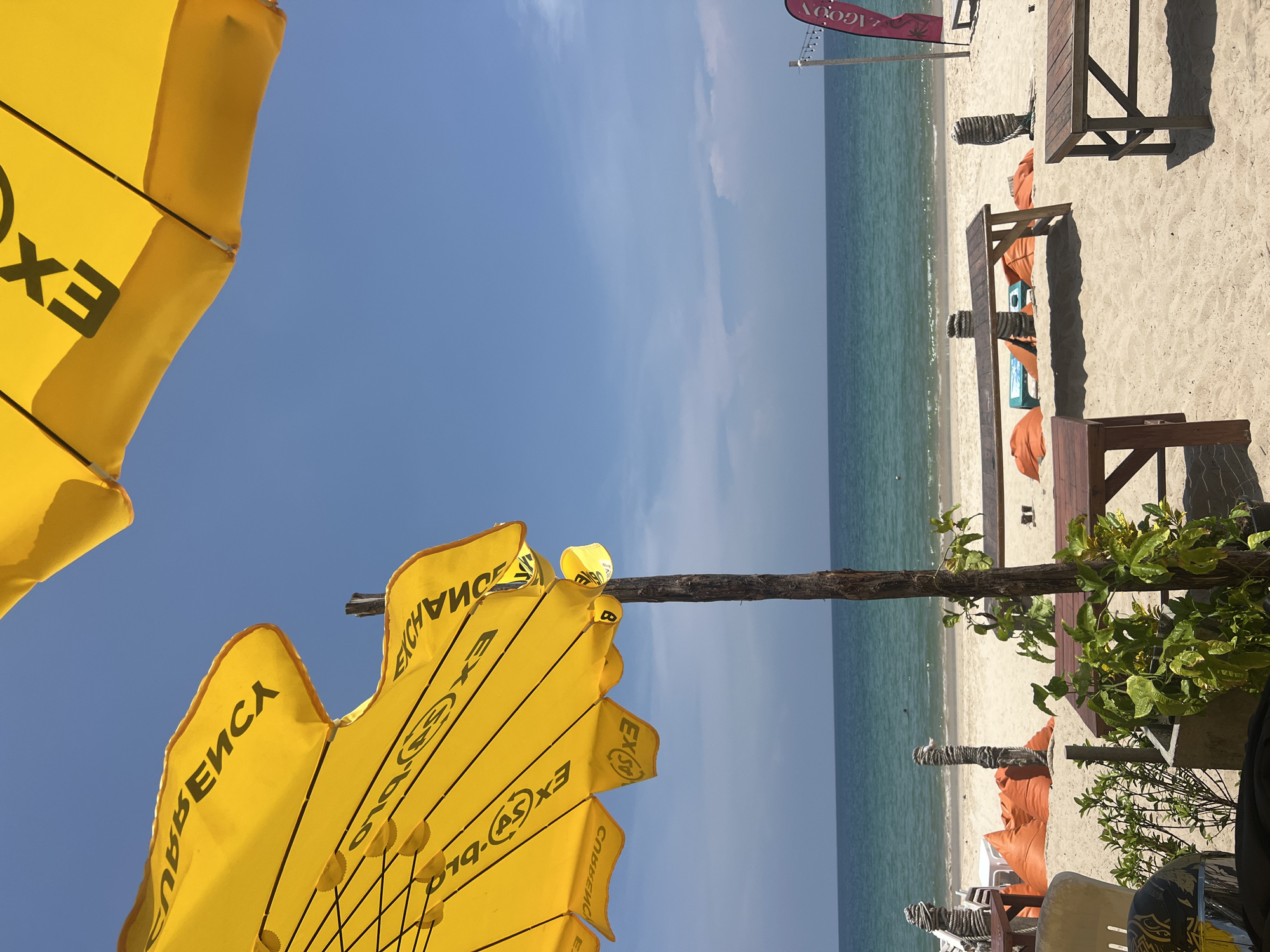 This image captures a vibrant, sunny beach scene, viewed from a slightly elevated position. In the foreground, two large, bright yellow beach umbrellas are prominent, partially obscuring the sky. The umbrellas feature black text and logos; visible words include "EXCHANGE", "EX", "GOLDE", and a partial Cyrillic word "ВЕЙСА".
Beyond the umbrellas, a wide expanse of sandy beach stretches out, meeting the calm, turquoise ocean under a clear, bright blue sky with a few faint clouds. Along the beachfront, several wooden tables and benches are set up on the sand, some paired with orange or peach-colored beanbag chairs, suggesting a seating area for a beach establishment. Potted green plants and a sturdy wooden post are also visible, contributing to the beach club aesthetic.
In the upper right corner, a red banner or flag can be seen, displaying the reversed text "LAGON" (likely "LAGOON"). The strong sunlight indicates a bright day, likely late morning or early afternoon, casting distinct shadows from the umbrellas and furniture. The scene appears peaceful and inviting, with no people visible, emphasizing the serene atmosphere of the beach.