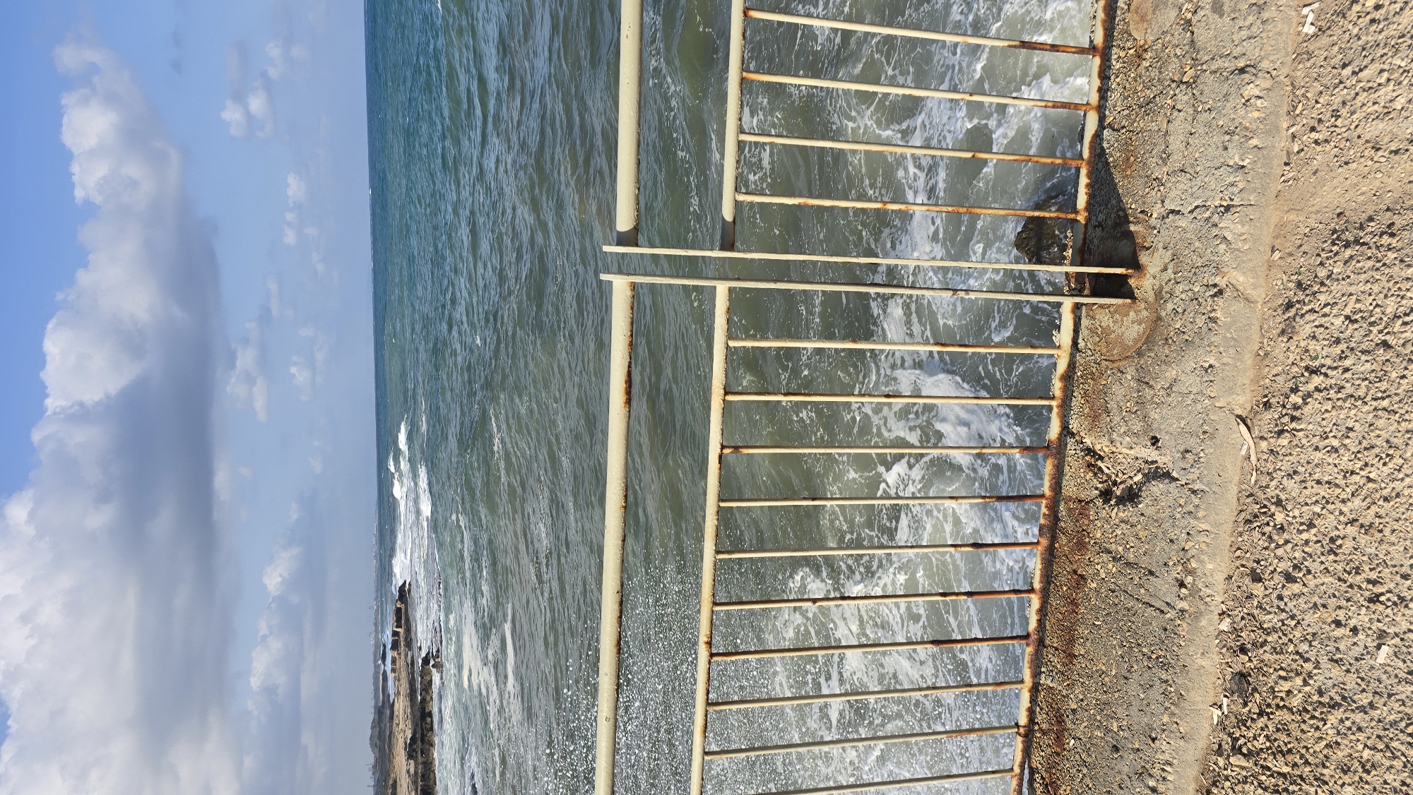 The image shows a view from a rusty metal fence looking out at the Mediterranean Sea, with waves crashing on a rocky shoreline. The sky is partly cloudy. The foreground consists of a gravelly path and a concrete embankment supporting the fence. The sea is a deep blue-green, with white foam from the waves. In the distance, along the coastline, there are some structures that appear to be part of a town or village. The location is identified as a coastal area in Mate Asher Regional Council, Israel. There are no people or animals visible in the image. The overall impression is one of a rugged and natural coastal environment.