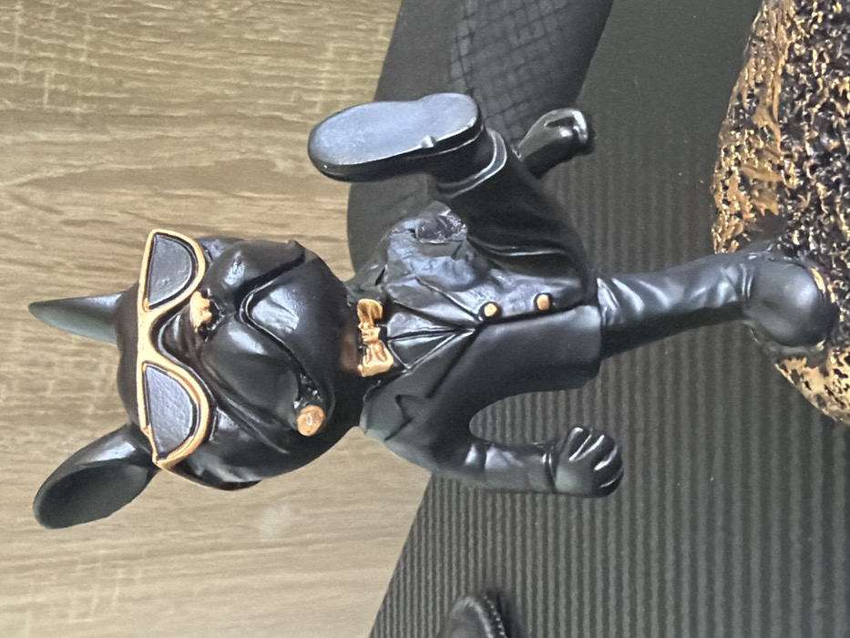 A black, anthropomorphic French Bulldog figurine, dressed in a black suit with gold buttons, a gold bow tie, and gold-rimmed dark sunglasses, is depicted mid-kick with its right leg raised, exposing the sole of its black shoe. A light brown cigar protrudes from its mouth. The figurine stands on an irregular, textured base with gold and dark brown coloration. The foreground consists of a dark, horizontally ribbed surface, while the background features a vertical wooden panel with a light brown grain. The image was captured in Sofia, Bulgaria.
