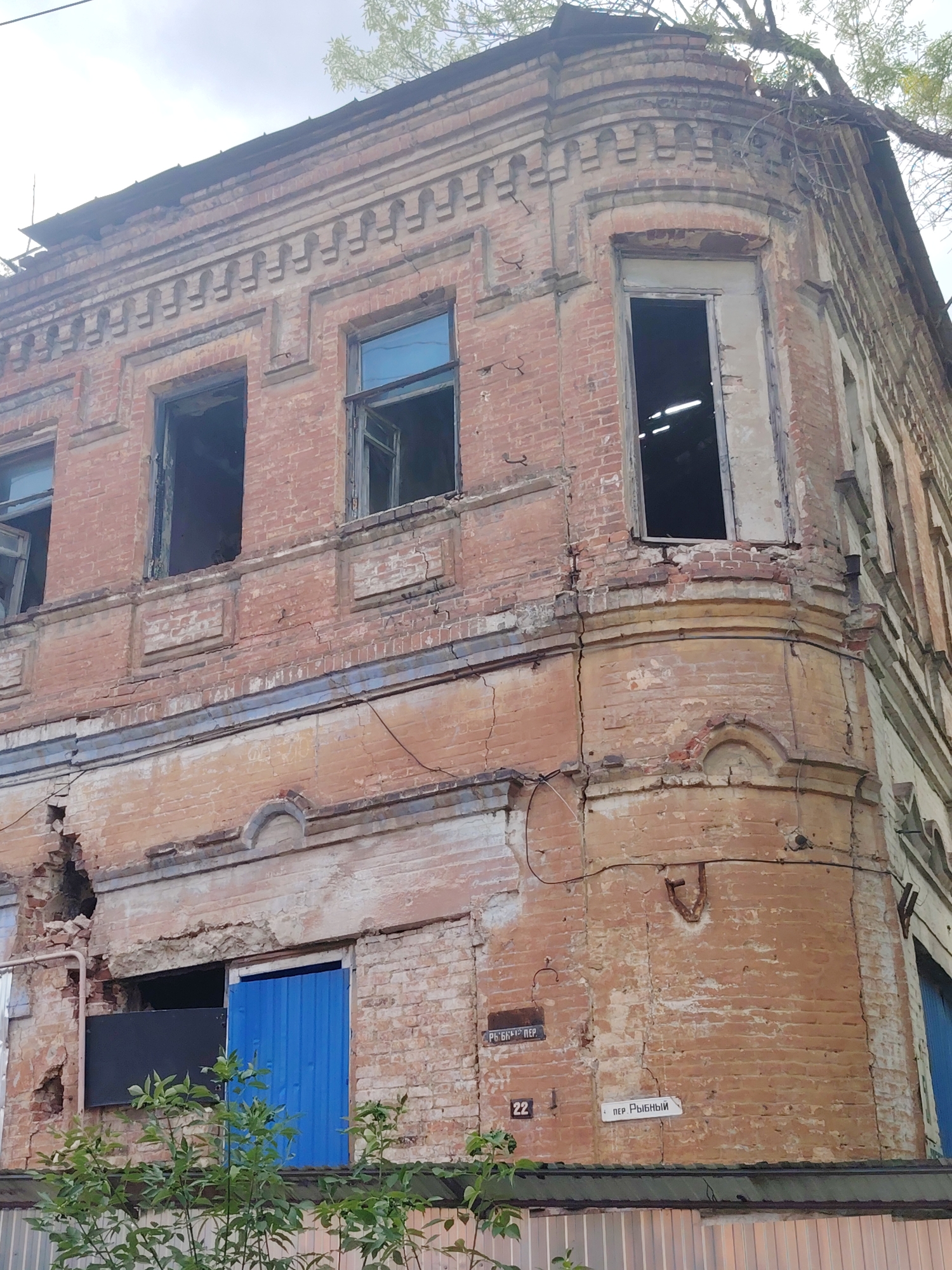 The image captures the exterior of a derelict brick building, likely an old apartment building, in Rostov-on-Don, Russia. The building shows significant signs of decay, with crumbling brickwork, cracked walls, and missing window panes. Some windows are boarded up, while others are gaping holes revealing the dark interior. A bright blue corrugated metal door has been installed in one of the ground-floor openings. The building's ornate architectural details, such as the decorative brickwork along the roofline and the curved corner, suggest it was once a substantial structure. Overgrown greenery, including a tree with branches extending over the building, further emphasizes its neglect.
Two street signs are visible on the building's facade. One, a white rectangular sign with black lettering, reads "Пер Рыбный" (Per Rybny), indicating a street name. Below it is a small square sign with the number "22". Another dark rectangular sign with white lettering, partially obscured by foliage, reads "РЫБКИН ПЕР" (Rybkin Per). The presence of these signs and the architectural style strongly suggest an urban or semi-urban setting in Russia.
The time of day appears to be daytime, with overcast skies suggesting potentially cloudy weather. The overall mood conveyed by the image is one of decay, abandonment, and forgotten history, contrasting with the vibrant blue of the newly installed door.