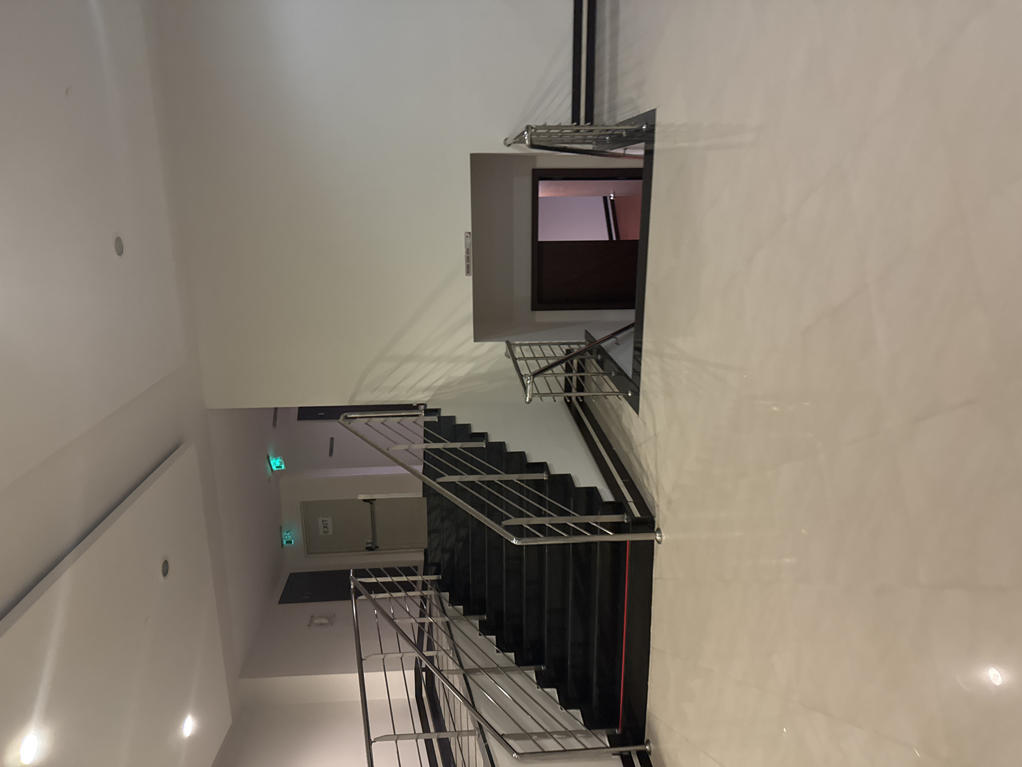 The image depicts an interior scene of a modern building, likely a hotel or office complex, located in Aksu, Turkey. In the foreground, a polished light-colored tiled floor reflects the overhead lighting. To the left, a dark-colored staircase with black steps and shiny metal railings ascends upwards. The staircase is bordered by a dark base.
Further into the scene, two green "EXIT" signs are visible on a wall. One is positioned above a door on the left side, and another is further back on the right. A closed door with a metallic handle is also present on the left. In the mid-ground, an open doorway leads down to a lower level, revealing a dark-colored door and a glimpse of a room with red flooring and pinkish walls. The overall environment is well-lit with recessed ceiling lights and presents a clean, contemporary aesthetic. There are no people visible in the image.