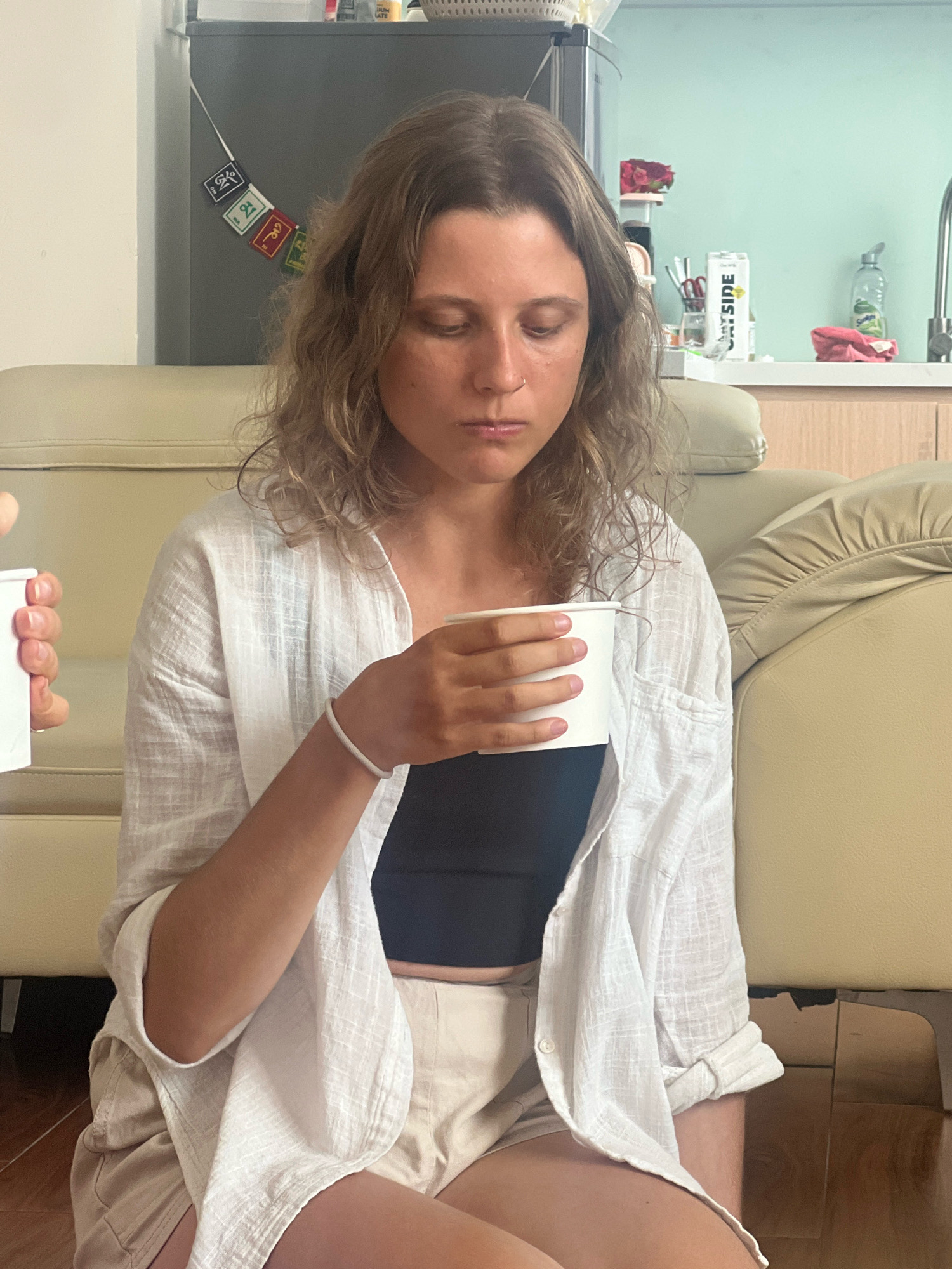 A young Caucasian woman with wavy blonde hair is seated on the floor, facing forward with her head slightly bowed, holding a white paper cup in her right hand. She is wearing a white, open linen shirt over a dark sleeveless top and beige shorts. A white bracelet is visible on her right wrist. The background features a cream-colored leather sofa, a refrigerator with small decorative flags, a kitchen counter with various items including a carton of "OATSIDE" oat milk, and a light blue wall. The scene appears to be indoors, in an apartment or house, likely in an urban setting such as the city of Đà Nẵng, Vietnam, suggested by the presence of the "OATSIDE" product which is popular in Southeast Asia.