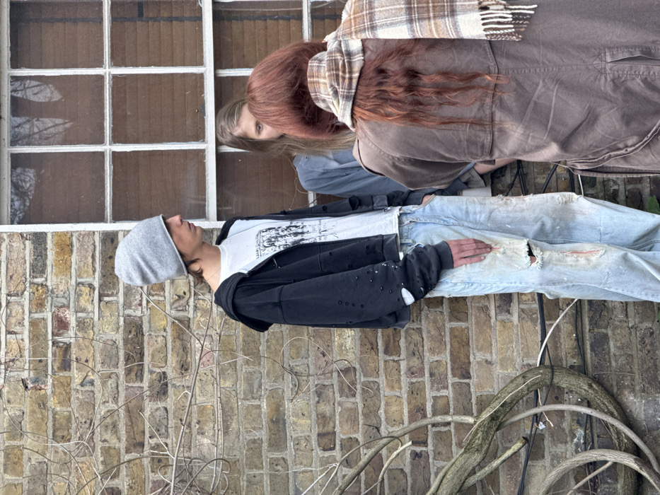 Three people are positioned against a building. A person wearing a grey beanie, white t-shirt, black jacket and torn jeans is lying against a brick wall. Two people are near a window. One is wearing a brown jacket and plaid scarf, and another with blond hair. A bare, woody vine curves in the foreground.