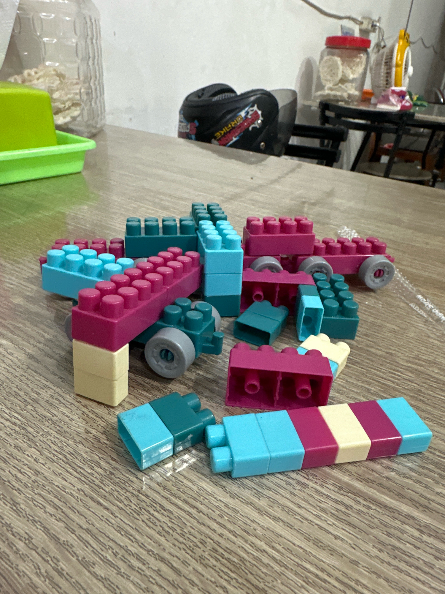 In a room in Purwotengah, Indonesia, a collection of colorful building blocks are scattered across a wooden table. The blocks, primarily in shades of blue, pink, and yellow, are arranged in various configurations, some stacked, others loosely placed. Among the blocks, a few have wheels, suggesting they are part of a larger construction or toy vehicle. In the background, partially visible, are a clear plastic bottle filled with white food items, a green plastic tray, a black helmet with colorful decals, and a small fan on a table. The overall environment appears to be a domestic setting, likely a home or playroom.