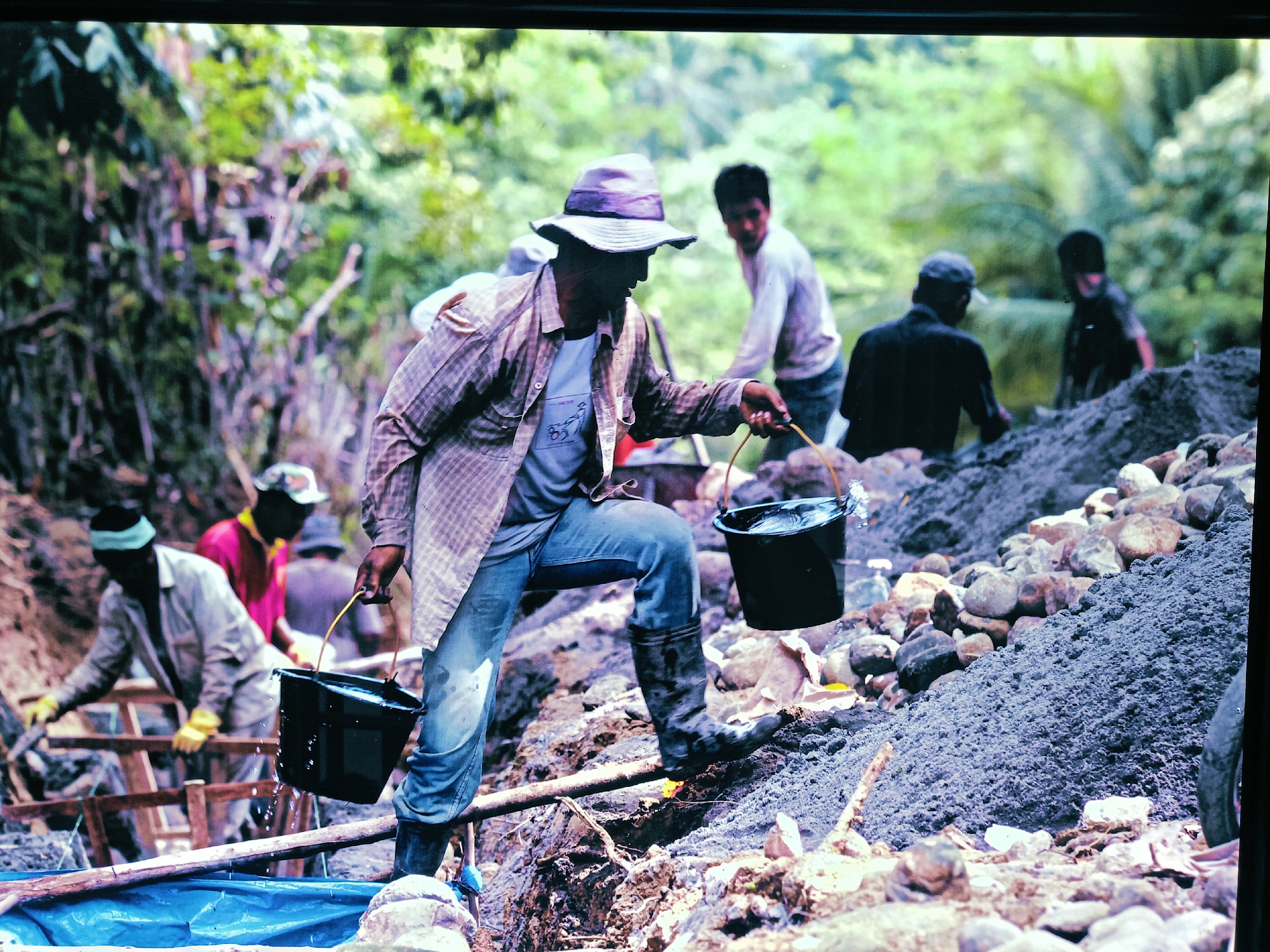 The image depicts a group of people engaged in manual labor in a landscape that appears to be in or near Banda Aceh, Indonesia.
A man in the foreground, wearing a hat, a plaid shirt over a t-shirt, jeans, and boots, carries two black buckets filled with liquid. He is positioned on a dirt and rock mound. Several other individuals are visible in the background. Some appear to be working with shovels and wooden structures. The environment consists of a terrain of rocks, dirt, and some greenery. The background features trees and possibly dense vegetation. Overall, the scene suggests manual labor, potentially related to construction or resource extraction.