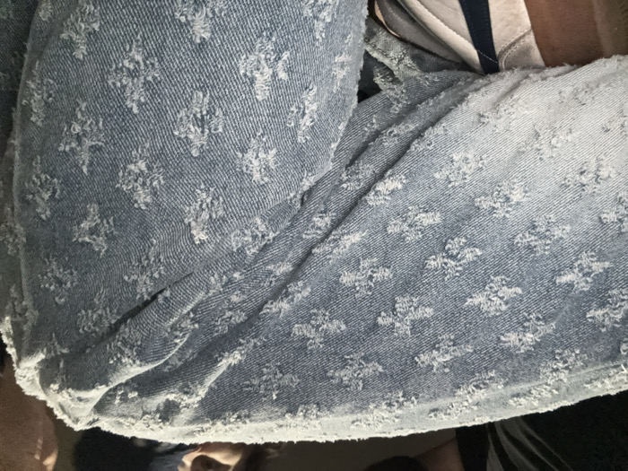 A close-up shot shows a light blue denim fabric, possibly a skirt or shorts. The material has a raised, textured pattern resembling small floral or star-like shapes. The fabric appears worn or distressed, with frayed edges adding to its casual aesthetic. A glimpse of clothing and a person's head and torso is visible.