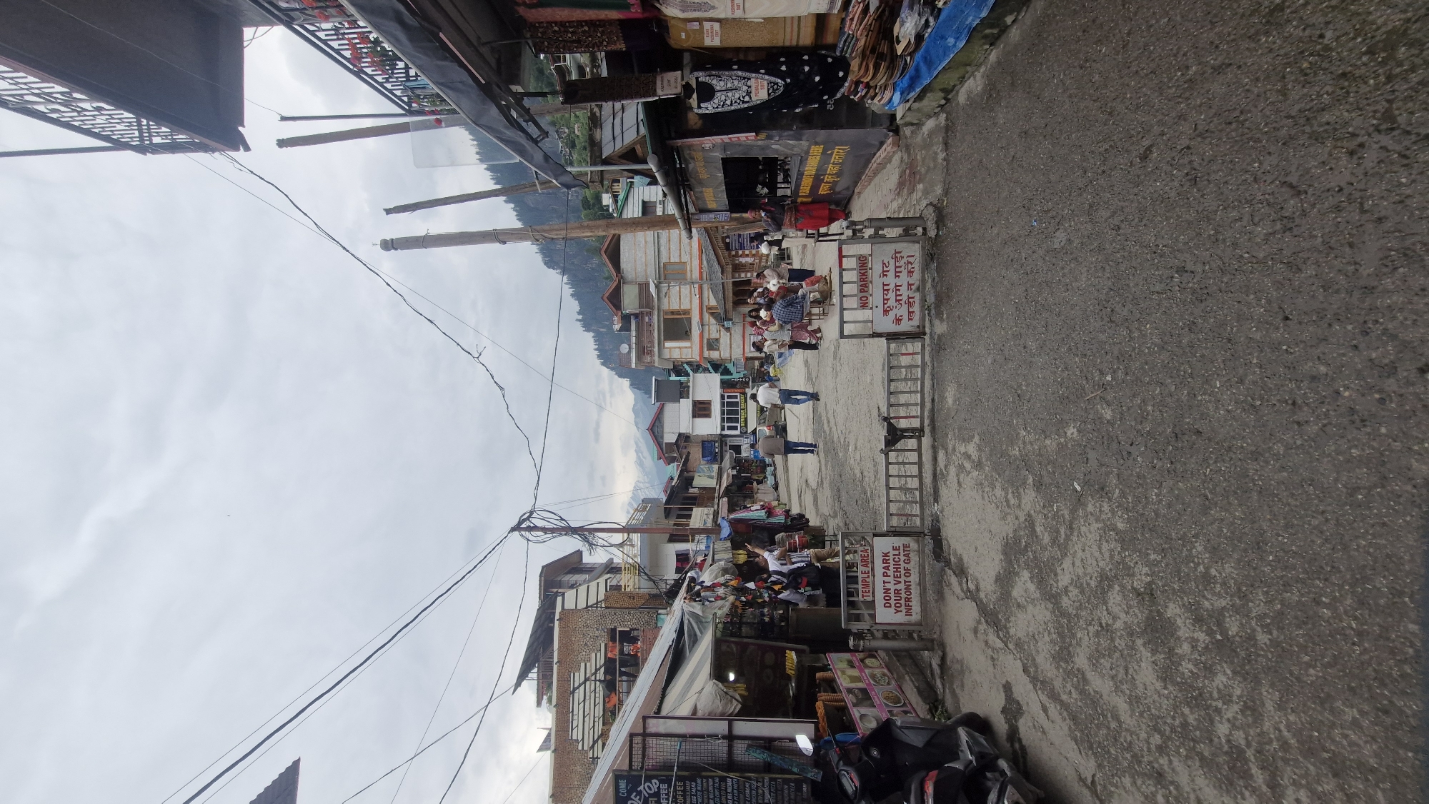 The image depicts a street scene in Vashisht, India, under an overcast sky. The street is paved and appears to be a commercial area with shops lining both sides. A metal gate with "NO PARKING" signs in English and Hindi is positioned in the middle of the street, blocking further passage.
On the left side of the street, several shops are visible. One shop has a menu board displaying items like "COFFEE," "OMELETTE," and "CHICKEN MEAT." A scooter is parked to the left of the gate.
On the right side of the street, more shops are present, some displaying clothing and textiles. People are seen browsing or standing near the shops. Two men in casual clothing are walking away from the camera down the street. In the distance, more buildings and the slopes of a mountain covered with trees can be observed. Telephone wires are strung across the sky above the street. The overall impression is that of a bustling marketplace in a mountainous region.