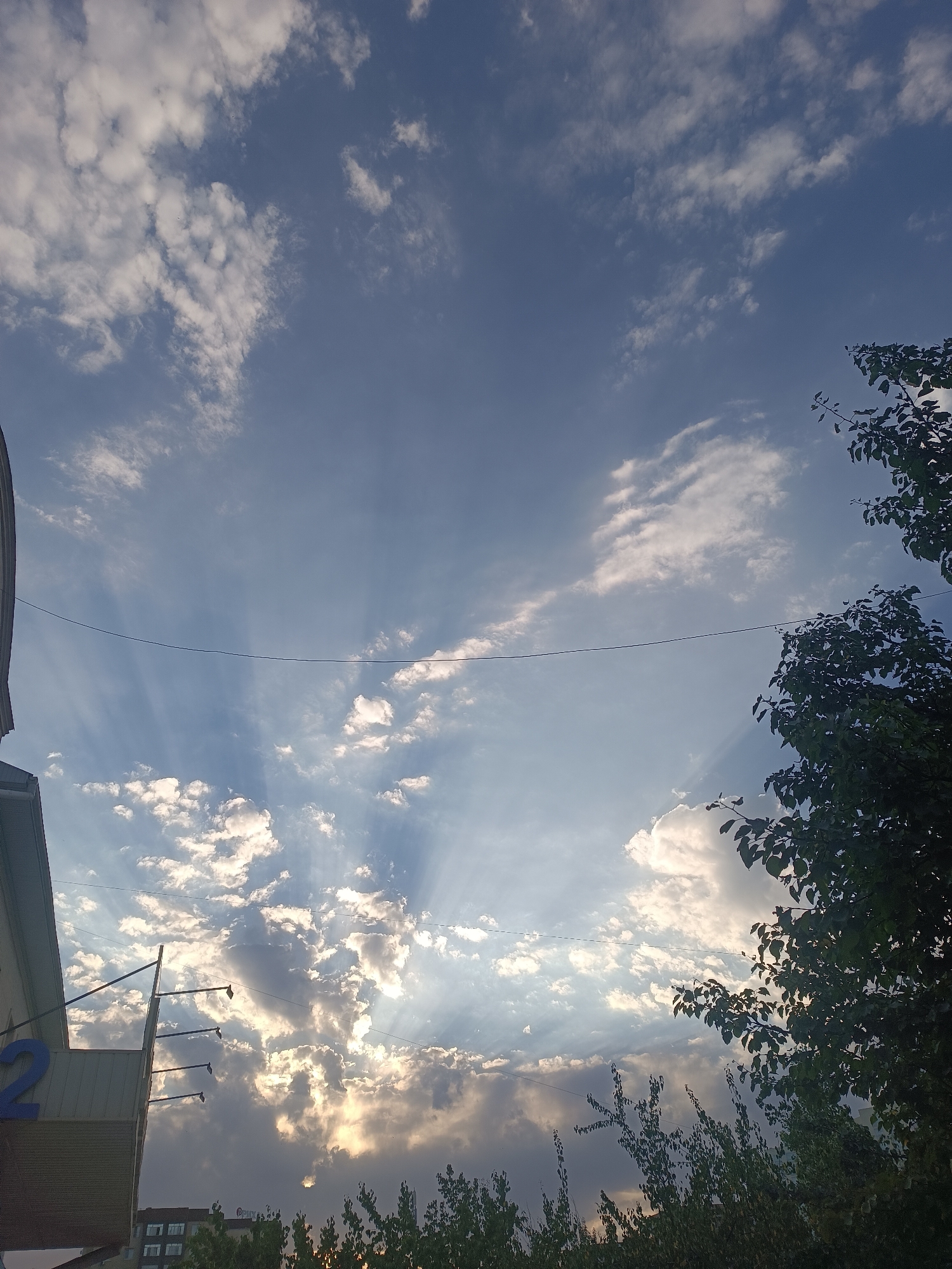 The image depicts a wide-angle view of the sky over Bishkek City, Kyrgyzstan. The sky is a dominant feature, filled with a mix of fluffy white clouds and thinner, wispy clouds, with rays of sunlight breaking through in several areas, creating a dramatic effect. The blue of the sky is deep and rich. In the foreground, on the left, a portion of a modern building is visible, featuring a large blue numeral "2" on its facade. Several dark poles with what appear to be light fixtures extend from the side of the building. On the right side of the image, a dense tree canopy with dark green leaves frames the sky. A thin, dark wire stretches horizontally across the middle of the frame, likely a power line or cable. In the lower part of the image, additional trees are visible, some appearing more distant and silhouetted against the bright sky. The overall scene suggests an outdoor environment, likely a street or open area within the city, during daylight hours, possibly late afternoon or early morning due to the lighting conditions. No people are visible in the image.