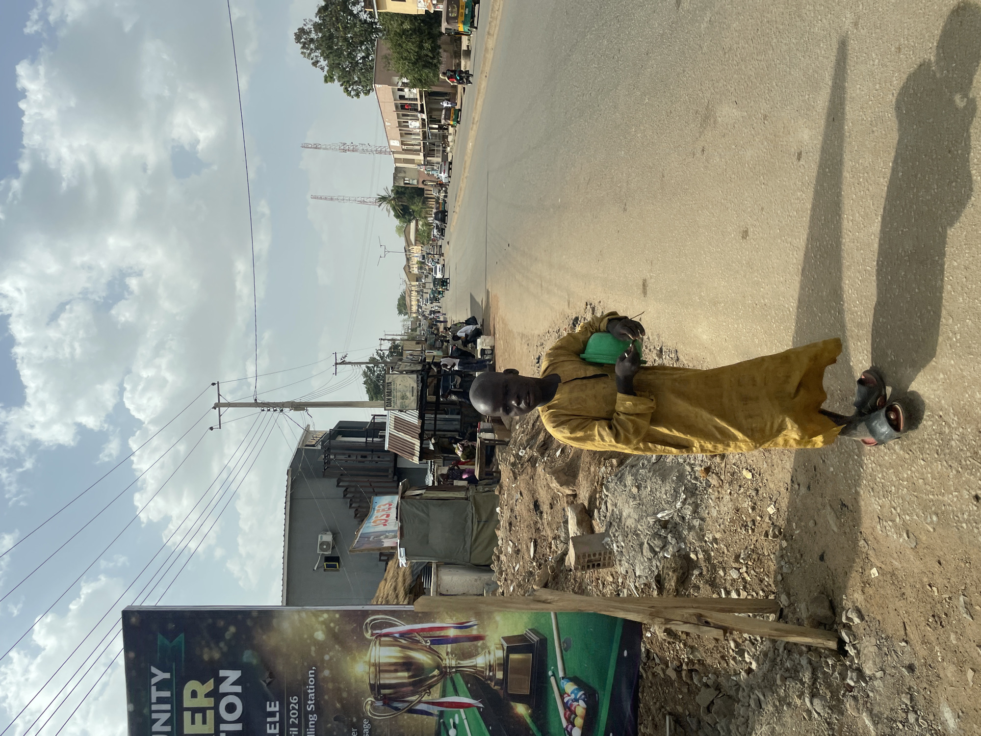 The image depicts a bustling street scene in Jos, Nigeria, under a bright, cloudy sky during the daytime. The sun casts distinct shadows, indicating its position.
In the foreground, a bald man of African descent is the central figure. He is dressed in a long, loose-fitting yellow tunic and dark sandals, and holds a small green object in his left hand. He stands on a dusty, rocky dirt shoulder beside an asphalt road, looking towards the camera with a neutral expression.
Next to the man is a large, colorful billboard, supported by two wooden posts. The billboard's design features a prominent golden trophy adorned with red, white, and blue ribbons, alongside a billiard table complete with cue balls and colored billiard balls. Visible text on the billboard includes "UNITY", "ER TION" (likely part of words like "Competition" or "Celebration"), "ril 2026" (suggesting a date in April 2026), "ding Station," (possibly "Filling Station" or "Trading Station"), and "ELE" (part of a location or event name).
The asphalt road stretches into the distance, with various vehicles, including cars and possibly a tricycle, present. The roadside is lined with numerous buildings, some appearing to be shops or residences, many featuring corrugated metal roofs. Overhead, power lines are strung between wooden utility poles. In the background, two tall, slender, red-and-white striped communication towers pierce the skyline. A small sign in the background also displays the text "DAN MAIYA". The overall scene portrays a typical urban or semi-urban street environment with ongoing daily activity.