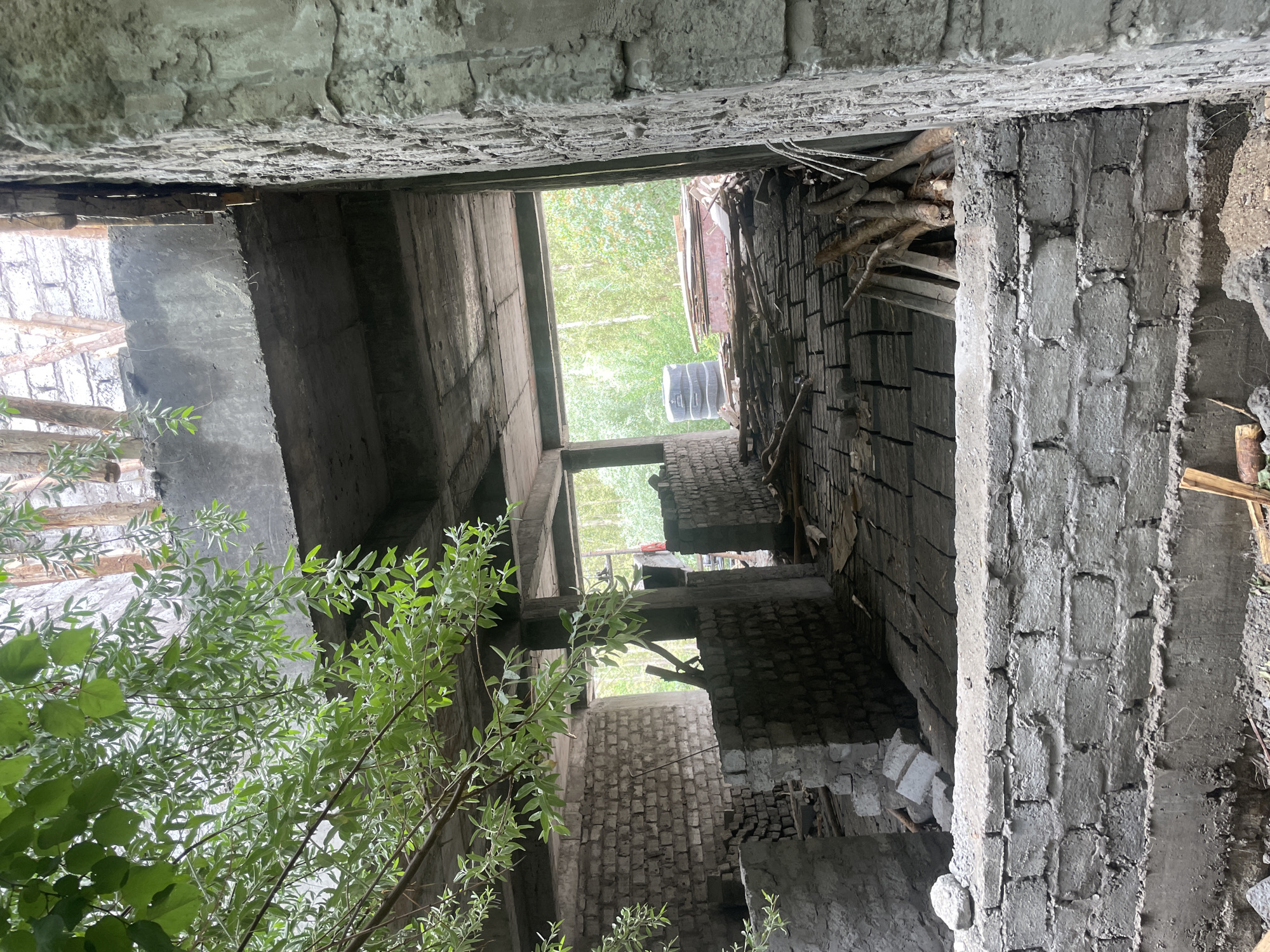 The image shows an interior view of an unfinished building, likely under construction, in the city of Leh, India. The structure appears to be made of concrete and brick. In the foreground, a low brick wall is visible, constructed with gray cement blocks. Behind this wall, the ground is covered with a dark, textured material, possibly bricks or pavers laid in a grid pattern, with some wooden logs scattered on top.
The interior walls are also constructed of brick. There are open doorways and unfinished concrete floors and ceilings, indicating that the building is in an early stage of construction. Sunlight streams in from an opening at the back, illuminating the interior and casting shadows. Outside the building, lush green trees and foliage are visible, suggesting a natural environment surrounding the construction site. The overall scene depicts a raw, unrefined construction environment.