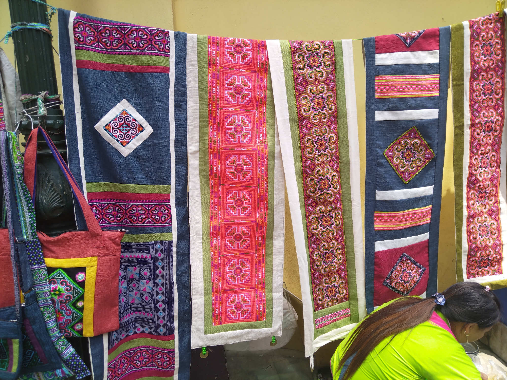 The scene depicts a display of textiles, likely for sale, in Sa Pa Ward, Vietnam. Several colorful fabric pieces with intricate embroidery hang side-by-side, including dark blue panels with diamond motifs, pink rectangular pieces with geometric patterns, and pieces with floral designs. A red tote bag with yellow and green accents hangs on the left. A person with dark hair and wearing a bright green top is partially visible on the right, looking downward. The background is a light yellow wall. A metal pole is visible on the left.
