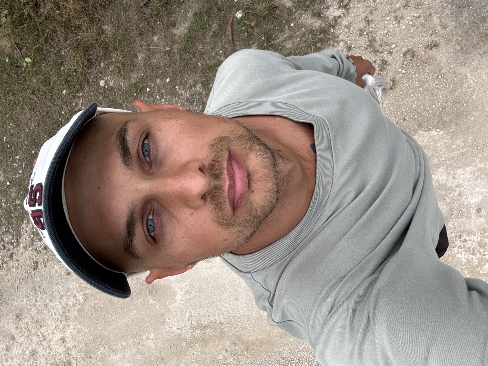 A man wearing a white baseball cap and a light grey long-sleeved shirt is standing outdoors. The cap has dark trim and a dark logo on the front. He has short hair, light-colored eyes, and a stubble beard. He is holding a clear plastic bag in his right hand. The ground around him is a mix of dirt, gravel, and dry grass. The background shows more of the natural environment. The overall scene suggests an outdoor location in a somewhat rural or undeveloped area, possibly a pathway or unpaved road within Kuta Selatan, Indonesia.