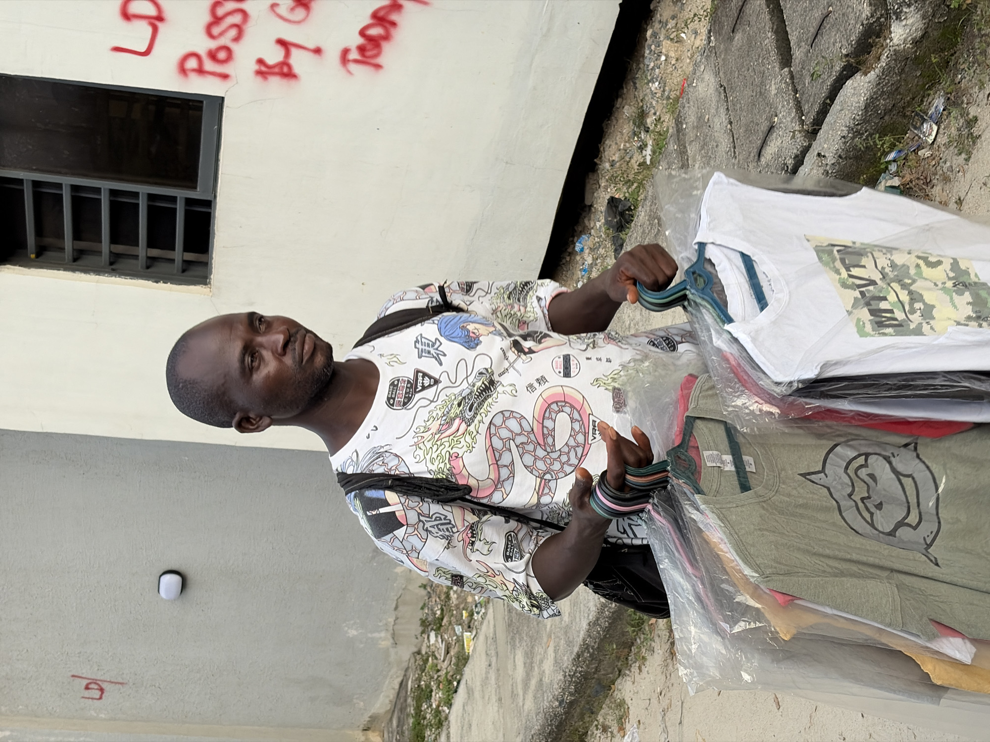 A street vendor is depicted in a brightly lit, vertical outdoor shot taken in Igboefon, Nigeria. The dark-skinned man, with a shaved head and a light beard, is positioned centrally, looking directly towards the viewer with a neutral, somewhat serious expression. He wears a distinctive white short-sleeved shirt patterned with colorful graphics, including a large dragon and Japanese-style imagery. A black bag strap crosses his chest.
In his hands, he holds a bundle of clothes hangers from which several new t-shirts are displayed, each individually sealed in clear plastic packaging. Visible t-shirts include a white one with a green and tan camouflage pattern and an olive-green one featuring a grey graphic of what appears to be an animal head. Colorful elastic bands are visible on his left wrist.
The background features a light-colored wall with a window fitted with dark metal bars. Red spray paint graffiti on the wall reads partially "LD Poss By G Tod". The ground is a rough mix of concrete and dirt, with scattered debris and sparse weeds, indicative of an urban or semi-urban street environment. The bright natural light suggests it is daytime.