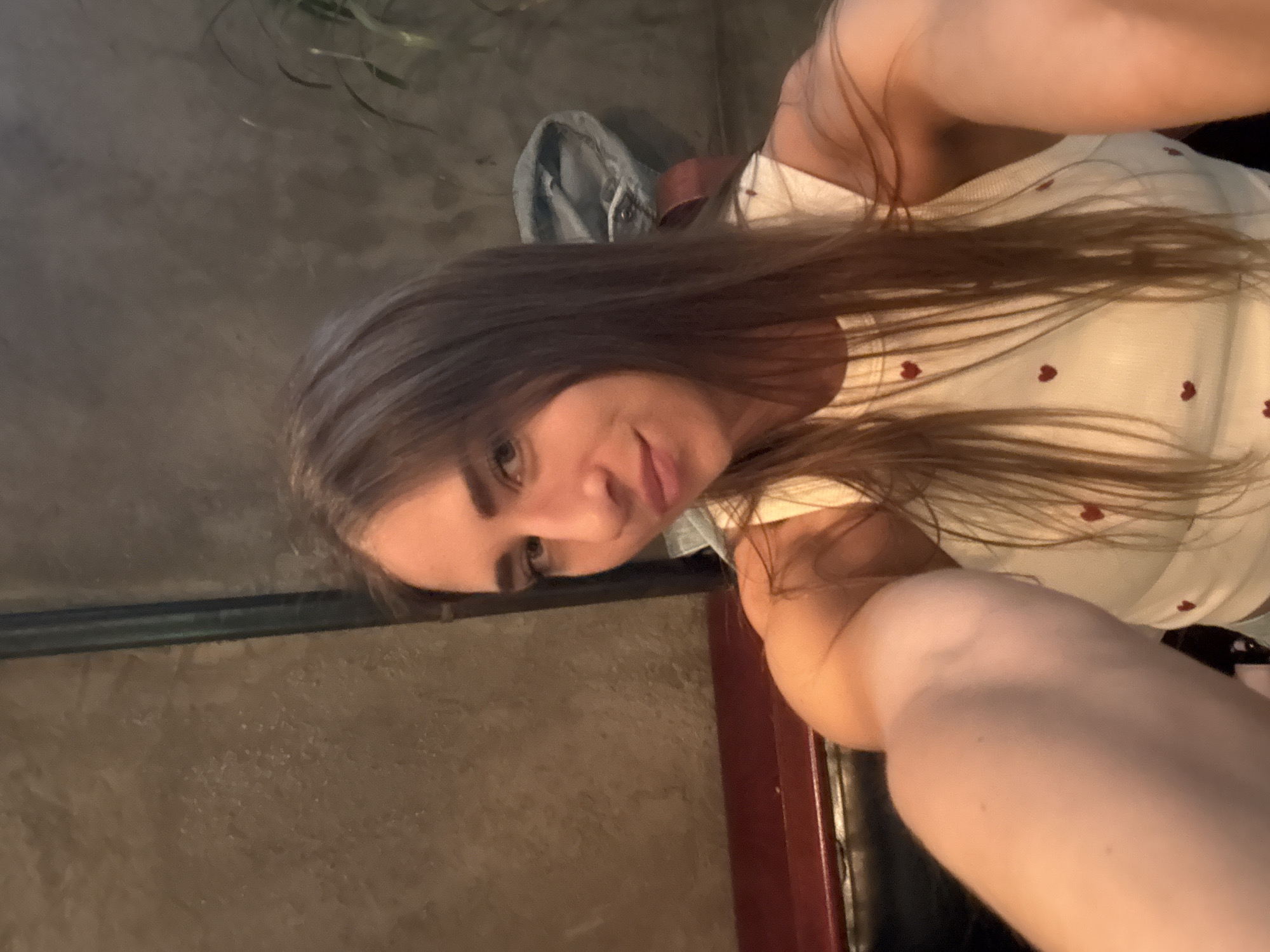 The image is a selfie of a young woman with long, brown hair. She is wearing a white tank top with small red hearts on it. Her face is partially visible, and she is looking at the camera with a slight smile. Her arm is extended towards the camera, holding it. The background appears to be an indoor setting with a concrete wall. There are no clear indicators of time of day or weather. The location context is Saint Petersburg, Russia.