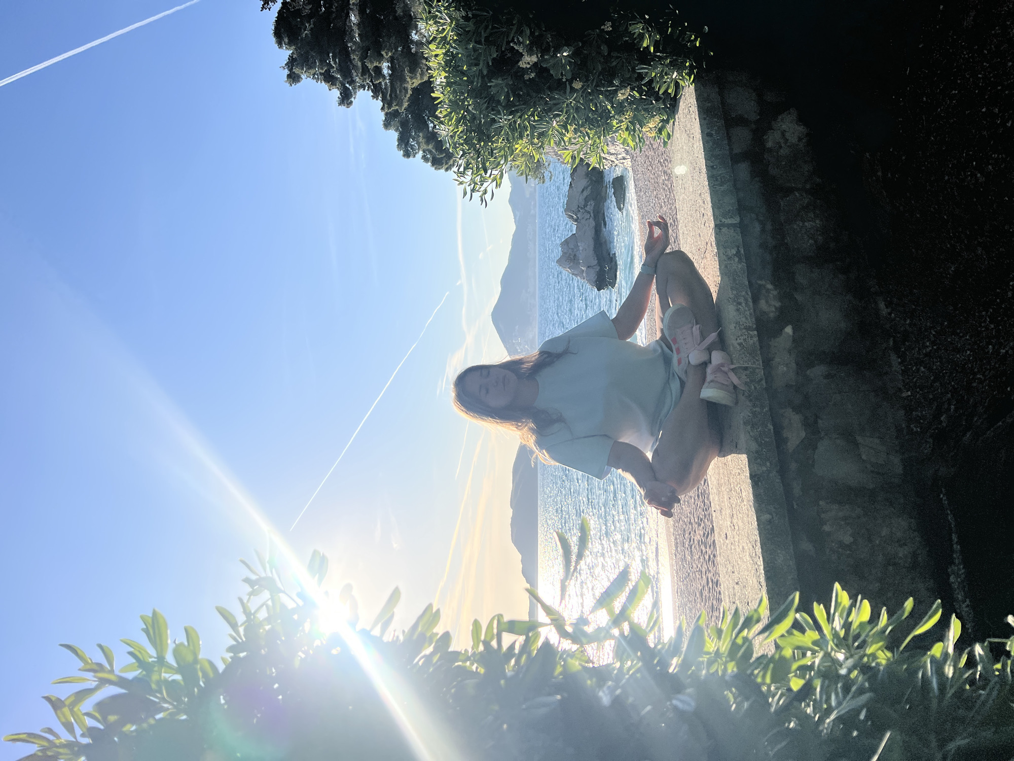 An outdoor, sunlit image depicts a young woman sitting on a stone wall overlooking a picturesque coastal scene, identified as Sveti Stefan, Montenegro. The woman, with long brown hair, wears a light blue t-shirt and light pink sandals. She is seated with her legs crossed, her left hand resting on her knee, and looks gently towards the viewer.
To her left, a calm, sparkling sea stretches towards distant, rugged mountains or cliffs under a clear blue sky. A prominent sun flare, indicative of either early morning or late afternoon light, emanates from the lower left of the frame (when correctly oriented), bathing the scene in warm, golden tones. A distinct white contrail from an aircraft streaks across the upper sky.
Lush green foliage frames the scene, visible in both the immediate foreground and as dense trees in the upper right background. The rustic stone wall provides a vantage point for the tranquil vista. The bright, clear weather and serene setting evoke a sense of peace and natural beauty. No visible text is present in the image.