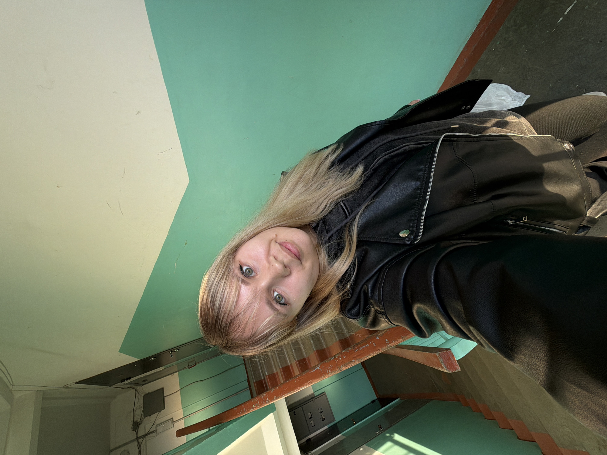 A young woman with long blonde hair and striking green eyes is featured in a selfie, taken from a low angle with a slight tilt. She looks directly at the camera with a gentle, pleasant smile. She is wearing a black leather jacket with silver zipper details, over a dark top, and dark trousers or leggings are partially visible.
The background depicts an indoor environment, likely a stairwell or hallway, characterized by its distinct two-toned walls. The upper part of the walls and the ceiling are painted a light cream or beige, while the lower section is a light teal or mint green, creating a sharp diagonal division across the frame. Below her, parts of a staircase are visible, featuring worn reddish-brown railings and textured or corrugated steps. In the deeper background, electrical wiring, conduits, and a grey utility box are discernible on the wall. A crumpled white plastic bag rests near her leg. The lighting creates clear highlights and shadows, illuminating the scene. No text is visible in the image.