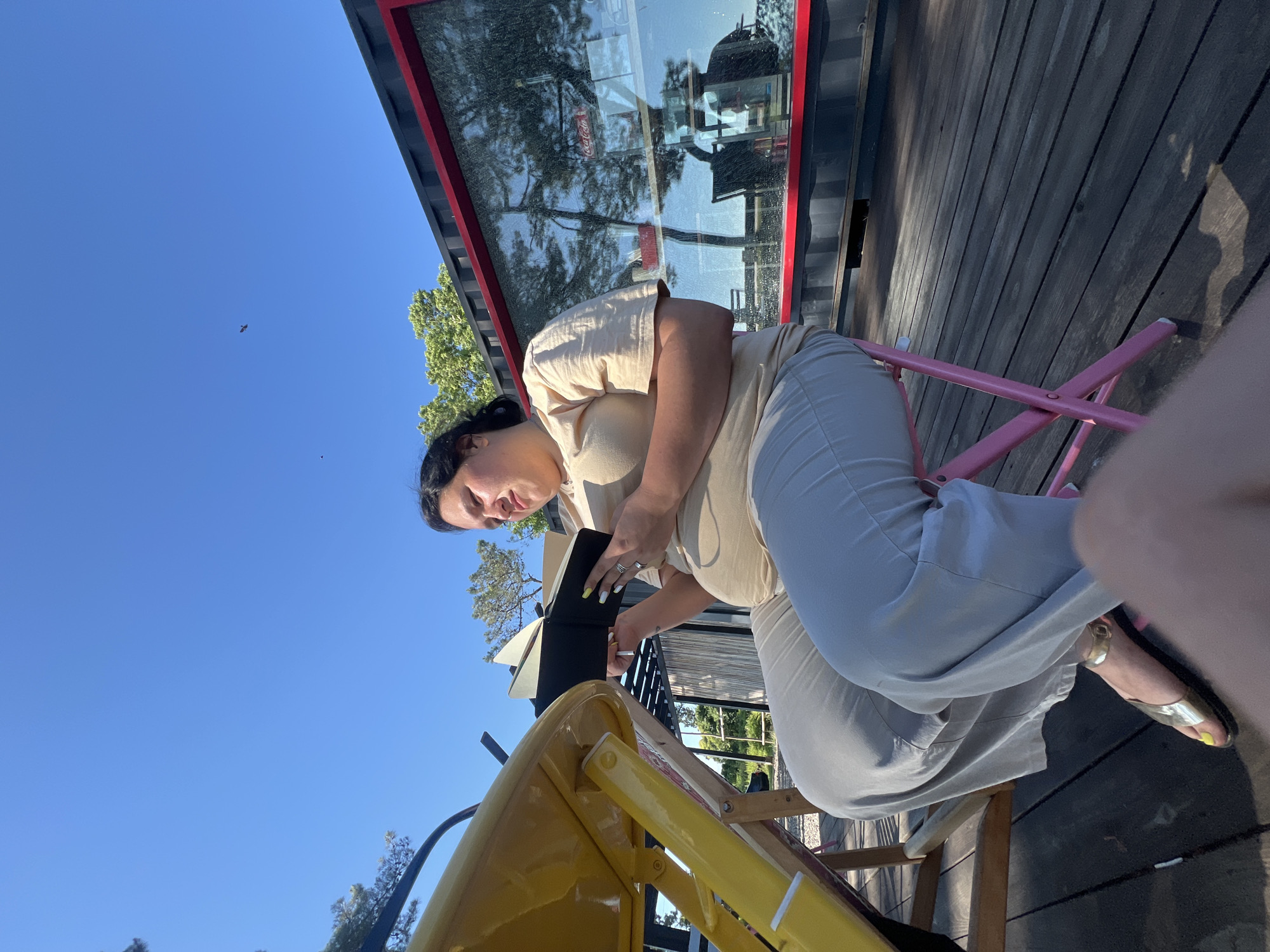 In this media file, a woman is captured from a slightly elevated perspective. She is seated on a pink chair, her body turned to her left as she looks down at a black notebook resting on a yellow easel. She holds a pen in her right hand, poised as if to write or draw, and her left hand is positioned on the notebook. She is wearing a beige, short-sleeved top and light-colored pants, with gold sandals on her feet. Her hair is dark and pulled back.
The setting appears to be outdoors, with a clear blue sky above and wooden planks forming the ground. Behind the woman, a building with a large glass window framed by a red border is visible. Reflections of trees and what appears to be a "Coca-Cola" logo can be seen in the glass. The presence of the easel and notebook suggests an activity like sketching or journaling. The lighting indicates it is daytime, likely sunny, given the clear sky and the shadows cast. The overall mood seems relaxed and focused, with the woman engrossed in her activity. The location context provided is Ochamchire, Abkhazia.