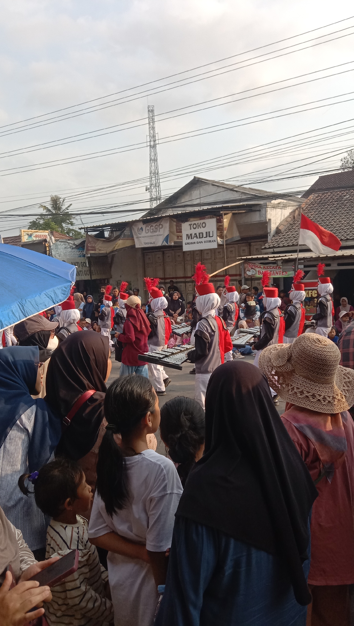A street parade is underway, with a marching band in uniform, featuring red feathered hats and white and red outfits, marching forward holding metallophones. Spectators are gathered on the side of the street, observing the procession. Some spectators are children, while others are adults wearing headscarves and hats. Buildings line the street, with signs visible, including "GGSP" and "TOKO MADJU". A red and white flag, likely the Indonesian flag, is prominently displayed. The sky is cloudy, and a communication tower is visible in the background. The overall scene suggests a community event or celebration in an urban or semi-urban setting.