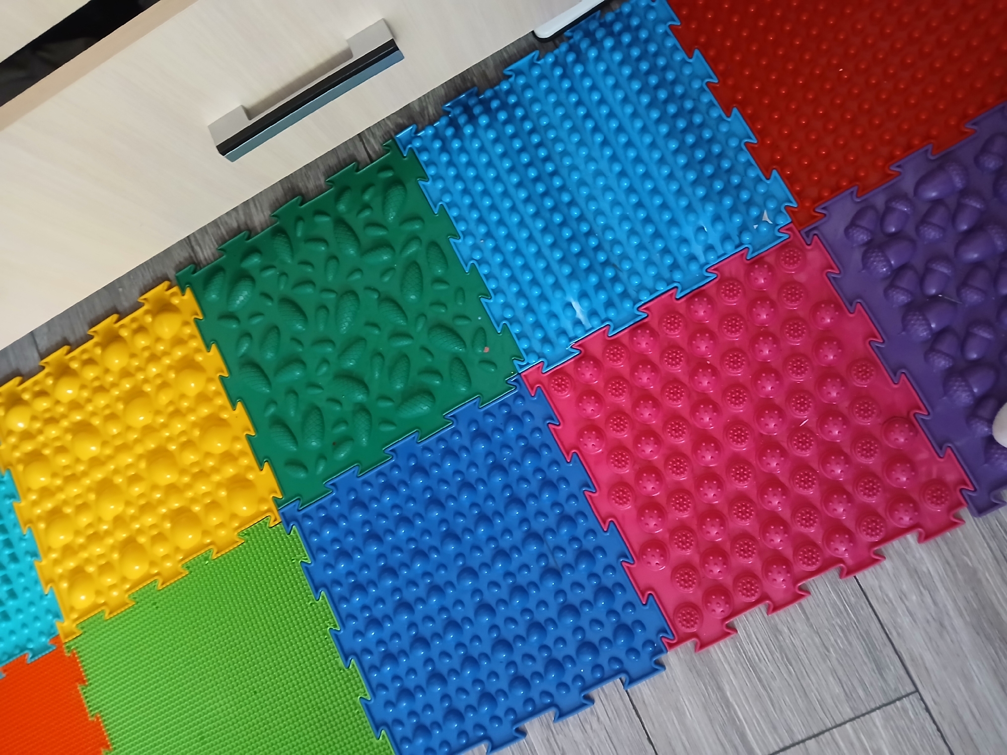 This is a close-up, overhead shot of a colorful, puzzle-piece floor mat arranged on a wooden floor. The mat is composed of several interlocking square tiles, each with a different textured surface and vibrant color. From left to right, the visible tiles are: orange, yellow, light blue, green, dark blue, red, pink, and purple. Some tiles have raised bumps, some have leaf-like shapes, and others have acorn-like shapes. The floor beneath is a light gray wood-grain laminate. In the upper left corner, a portion of a light-colored wooden cabinet with a dark handle is visible. The lighting suggests an indoor setting, likely during the day. There is no text visible in the image. The scene appears to be a playroom or exercise area, possibly for children, given the bright colors and the nature of the mat, which is often used for sensory play and foot massage. The focus is entirely on the textured mat and its arrangement, creating a visually dynamic and playful composition.