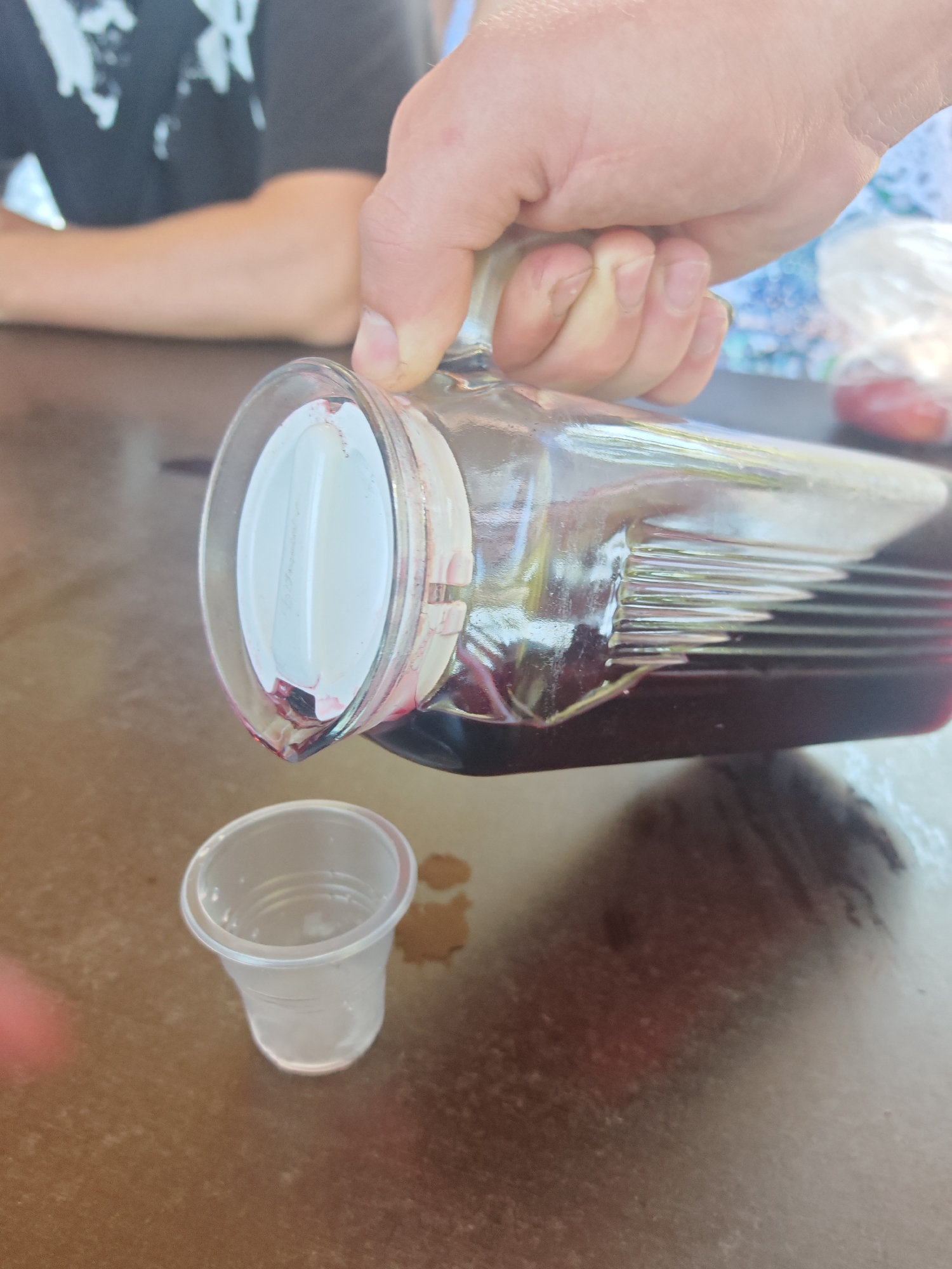 In Varvarovka, Russia, a hand is pouring dark red liquid from a glass pitcher into a small clear plastic cup. The pitcher has a white lid and visible ridges along its side. The scene takes place on a brown, possibly wooden, table. In the background, a person wearing a black t-shirt is partially visible, along with other out-of-focus elements that suggest an outdoor or casual setting.