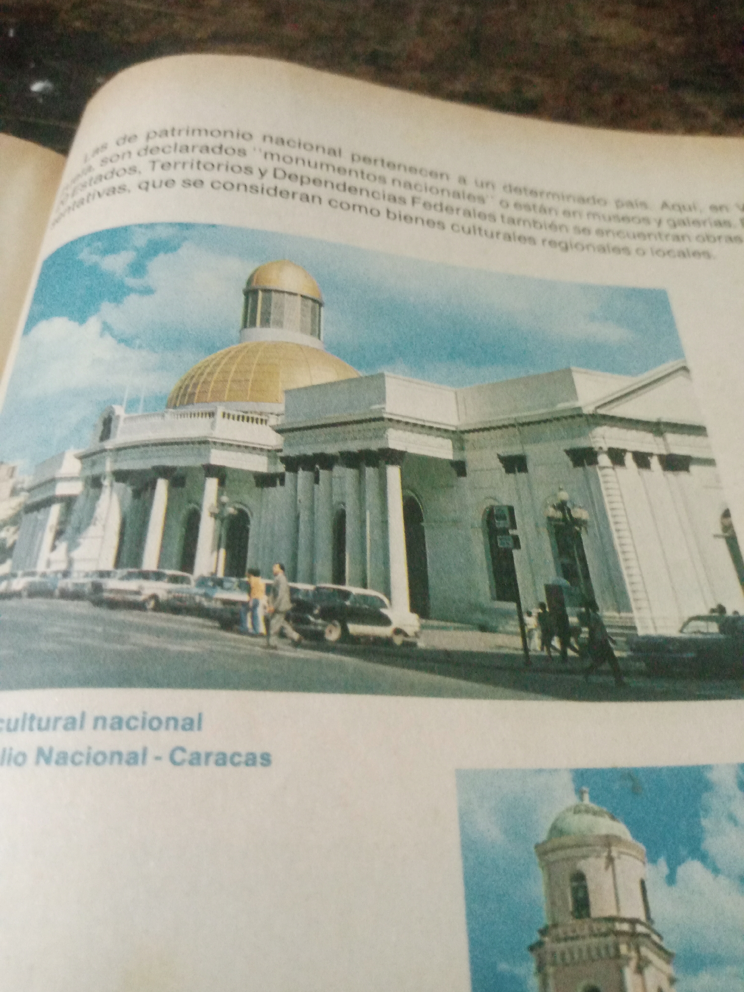 The image displays a historical building with a large golden dome, characteristic of public architecture. The building features classical columns and a grand facade. Several vintage cars are parked along the street in front of it, suggesting it might be a significant landmark. Two people are walking on the sidewalk, one in a light-colored suit and the other in casual attire.
Beneath this main image, there is text indicating "cultural nacional" and "lio Nacional - Caracas". This suggests that the main image is of a national cultural site located in Caracas, Venezuela, specifically the Capitolio Nacional. The image also shows a smaller picture of a bell tower with a green dome, also against a blue sky with clouds. The style of both buildings and the cars suggests the photos were taken sometime in the mid to late 20th century. The location of Santa Ana, Venezuela, is not depicted in the provided image.