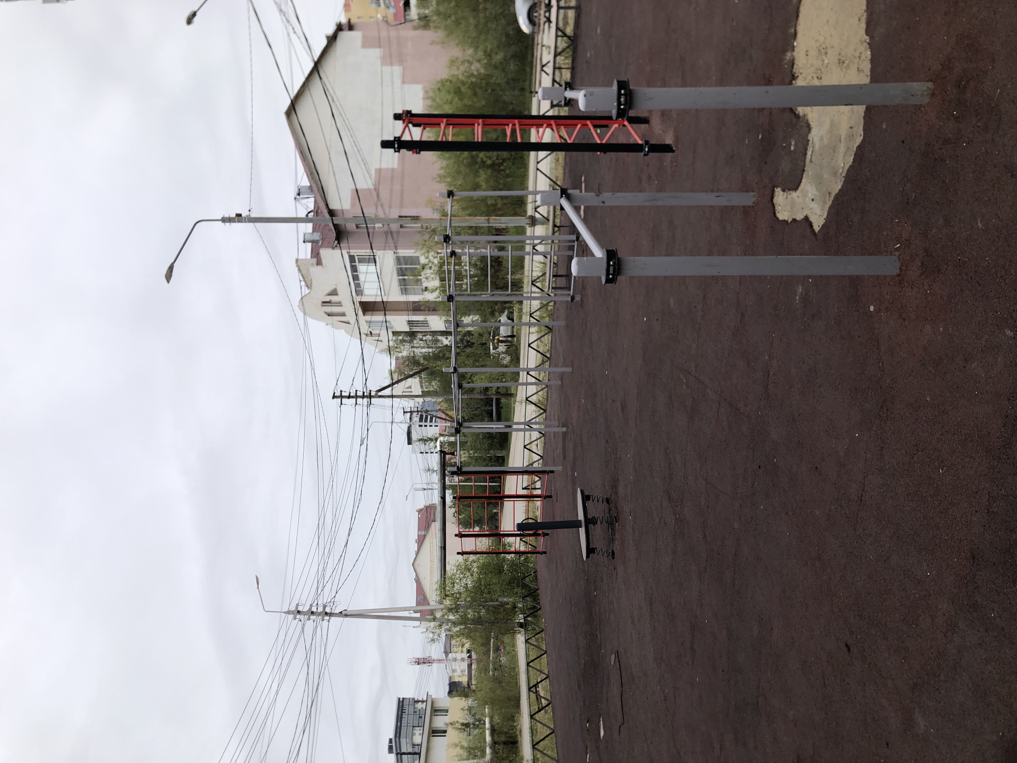The image depicts an outdoor sports ground in Yakutsk, Russia, on an overcast day. The ground is covered with dark, impact-absorbing material. Several pieces of exercise equipment are present, including parallel bars, a ladder, and a single-person spring rider. A low, triangular fence borders the sports ground. In the background, buildings of various architectural styles are visible, some with pitched roofs and others with flat roofs. Overhead, a complex network of power lines stretches across the sky, supported by utility poles. The overall impression is of a functional, if somewhat utilitarian, urban public space.