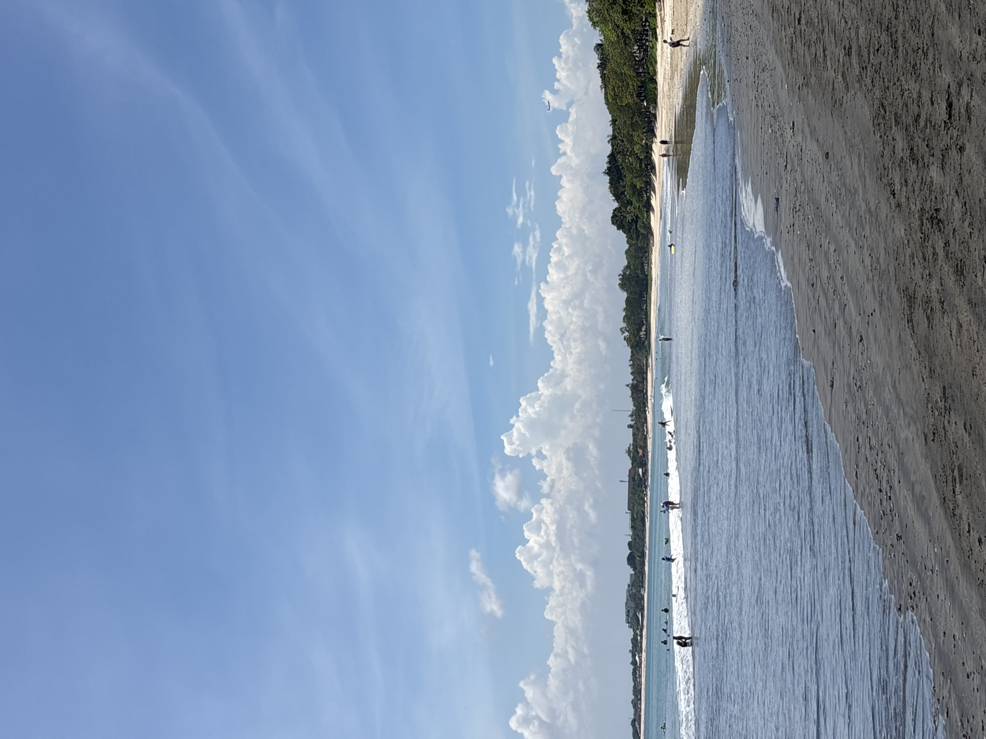 The image depicts a beach scene in Kuta Selatan, Indonesia. The sky is predominantly blue with some wispy clouds and a line of larger, cumulus clouds near the horizon. The beach stretches across the right side of the image, featuring tan sand that transitions into the ocean. Small waves break near the shore. There are people in the water, some possibly surfing, and a few individuals walking along the beach. A line of trees and vegetation borders the beach, creating a green backdrop between the sand and the clouds.
