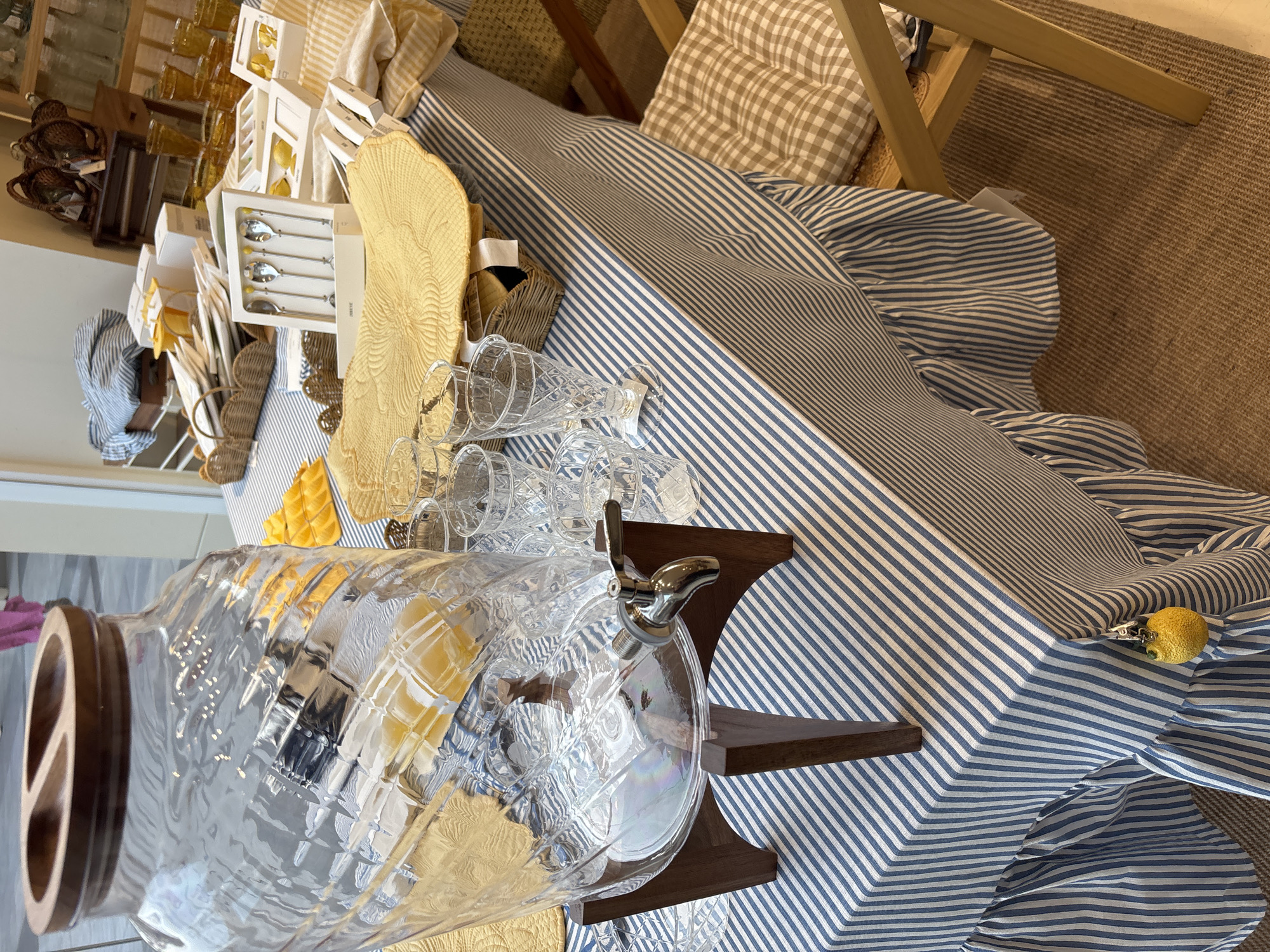 The image depicts a tablescape likely set up for a beverage service, possibly in a retail or hospitality setting. A large, clear glass beverage dispenser with a wooden lid and a metal spigot is prominent on the left side of the frame, filled with a light-colored liquid, likely juice. The dispenser rests on a dark wooden stand. The table is covered with a blue and white striped tablecloth that has a ruffled hem.
Several clear glass tumblers of various sizes and designs are arranged on the table. In the background, to the right of the dispenser, are more glasses, some appearing to be amber-colored.
Scattered on the table are various items, including yellow placemats with a woven texture, and several white boxes containing what appear to be cutlery sets and possibly other tableware, one of which prominently displays four spoons. A small, bright yellow lemon charm is hanging from the ruffled edge of the tablecloth on the right side of the image.
The setting includes a wooden chair with a checkered cushion to the right of the table. In the far background, there are shelves filled with bottles and woven baskets, and a glimpse of a person wearing a pink garment. The overall ambiance suggests a setup for serving drinks, possibly with a summery or picnic theme due to the yellow accents and lemon charm. The location is identified as Orihuela, Spain.